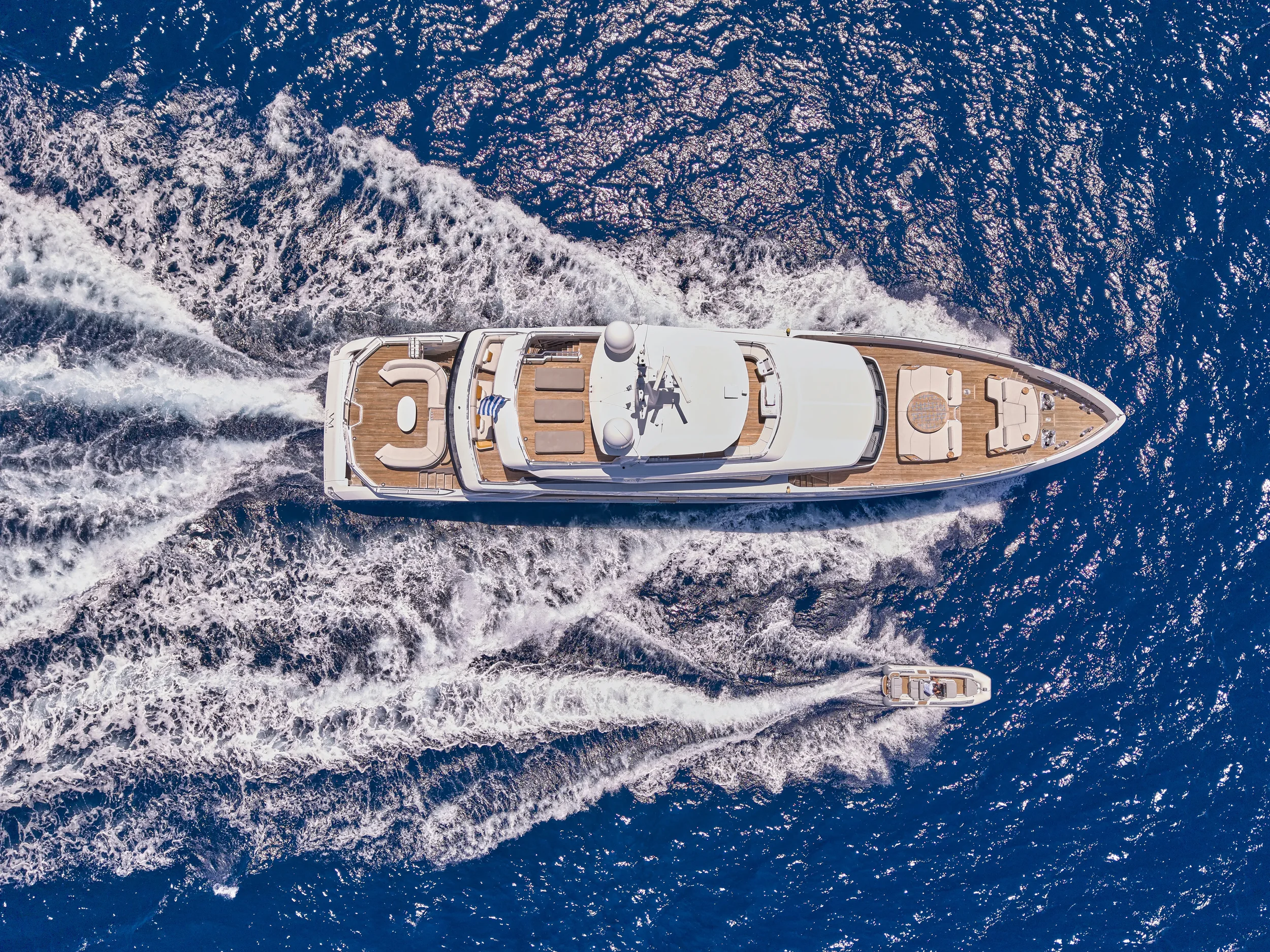 An aerial view of a white yacht with a smaller boat following in open blue water.