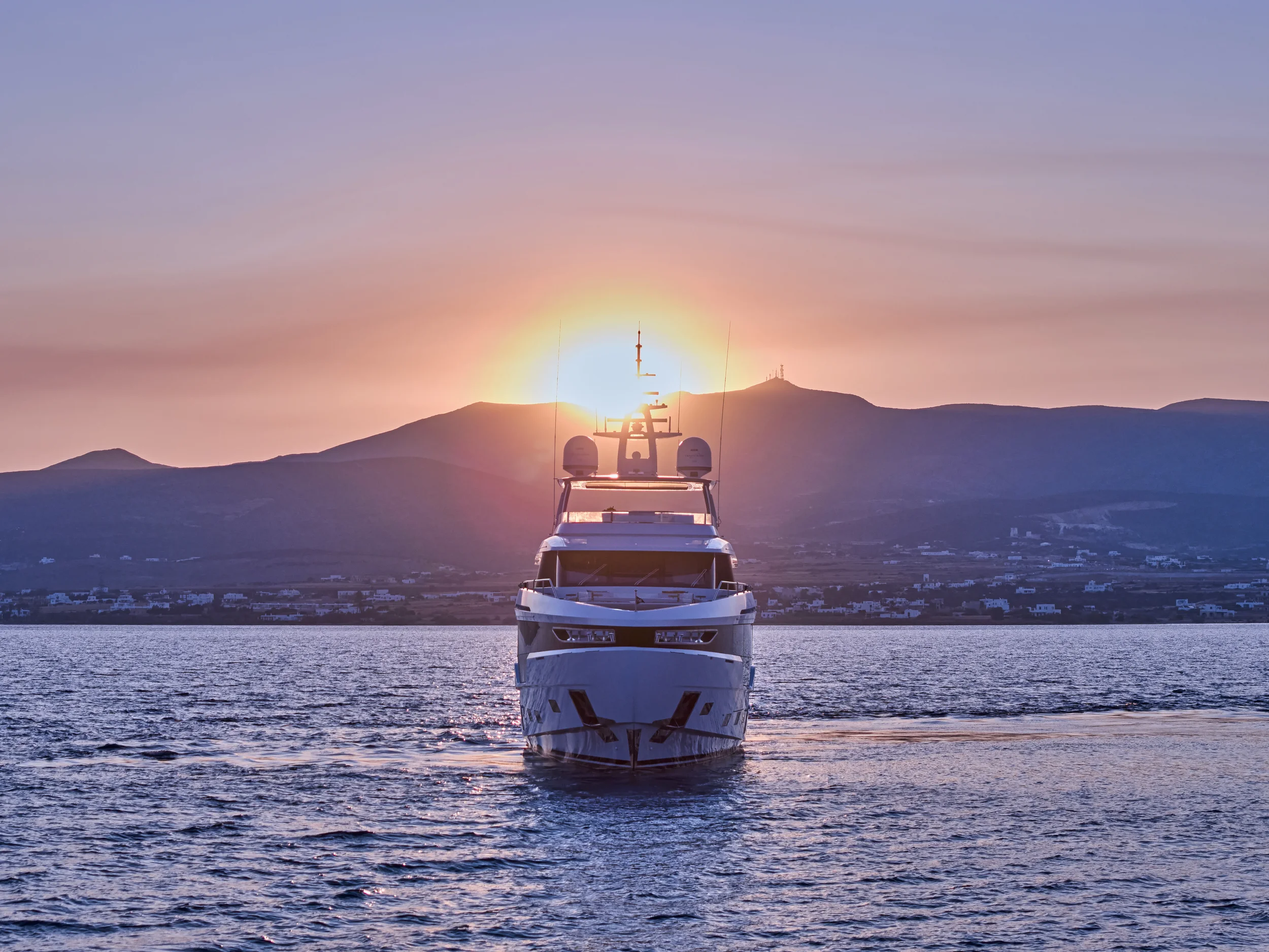 Luxury yacht sailing on calm water at sunset with mountains in the background.