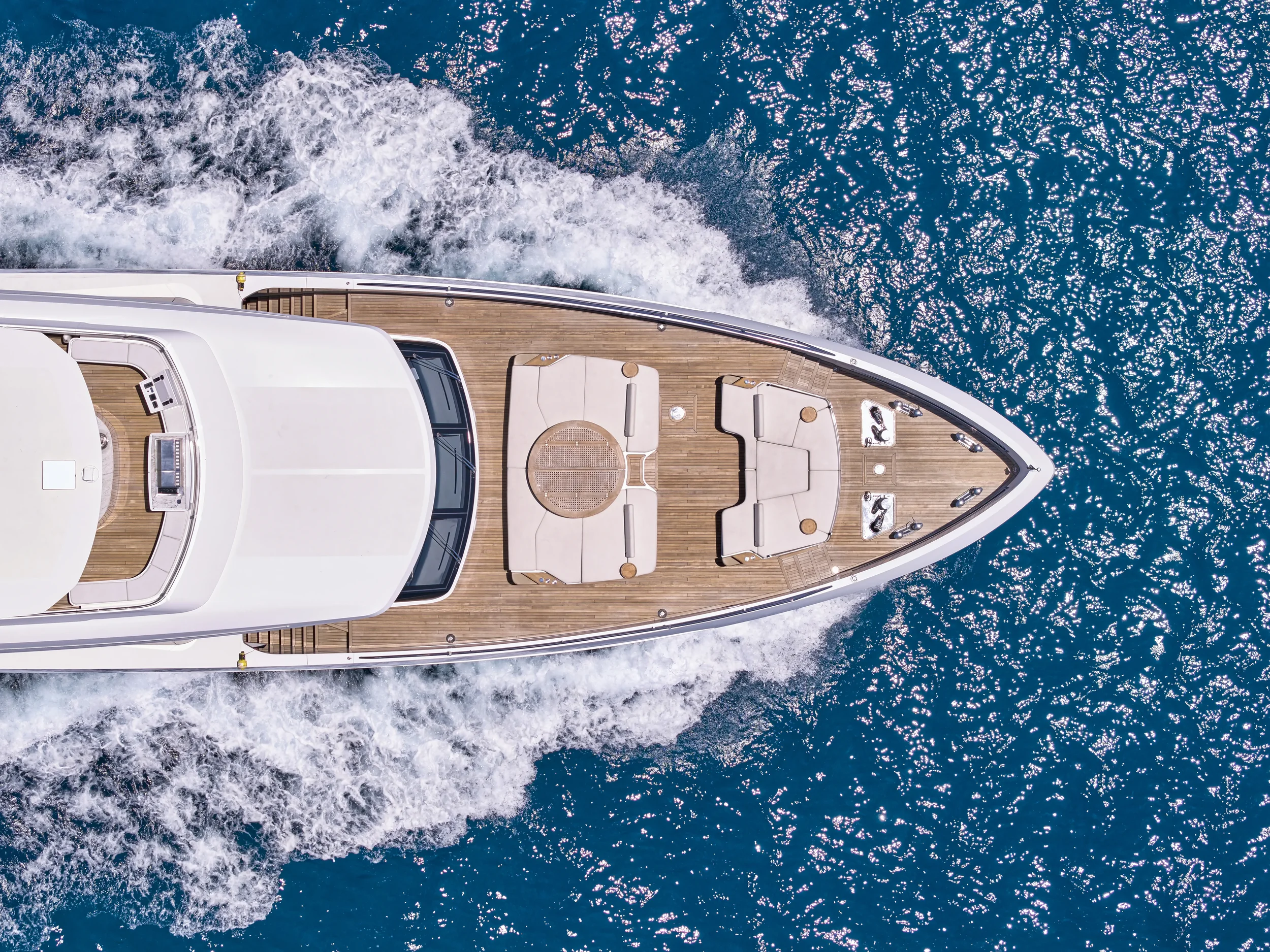 Top-down view of a luxury yacht sailing on the ocean, showing a wooden deck with seating areas and a small table, surrounded by blue water with white foam from the boat's movement.