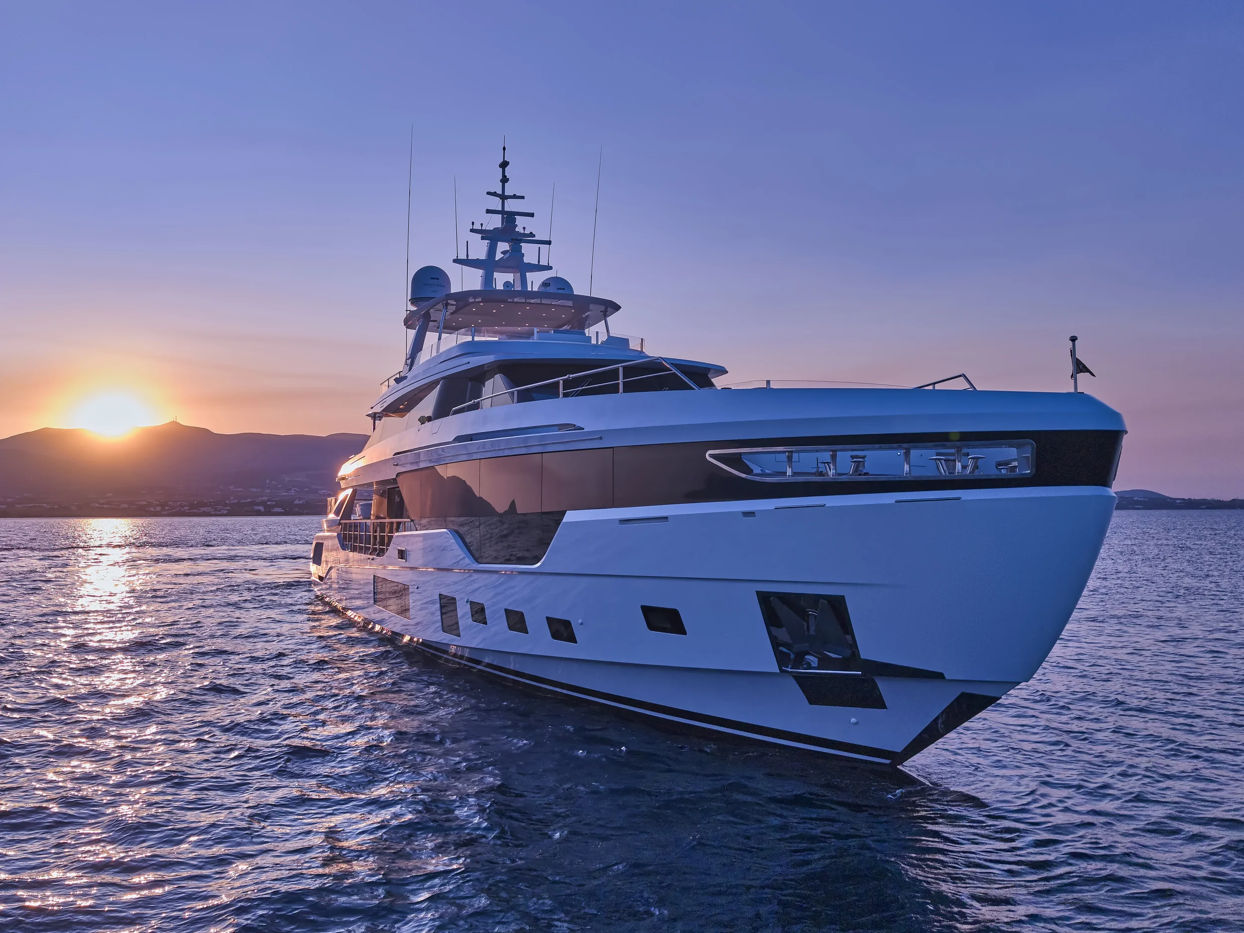 A large white yacht sailing on calm water during sunset with hills and a partly cloudy sky in the background.