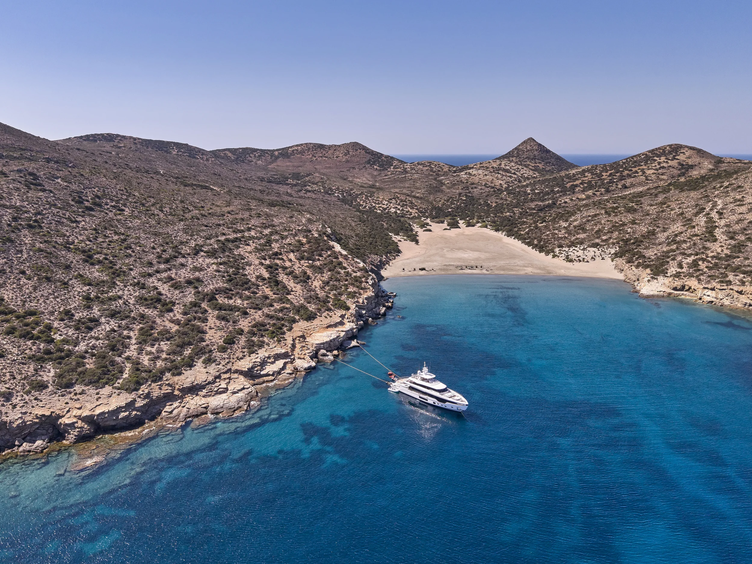 A white yacht anchored in a small bay with clear blue water, surrounded by rocky hills with sparse vegetation, under a clear sky.