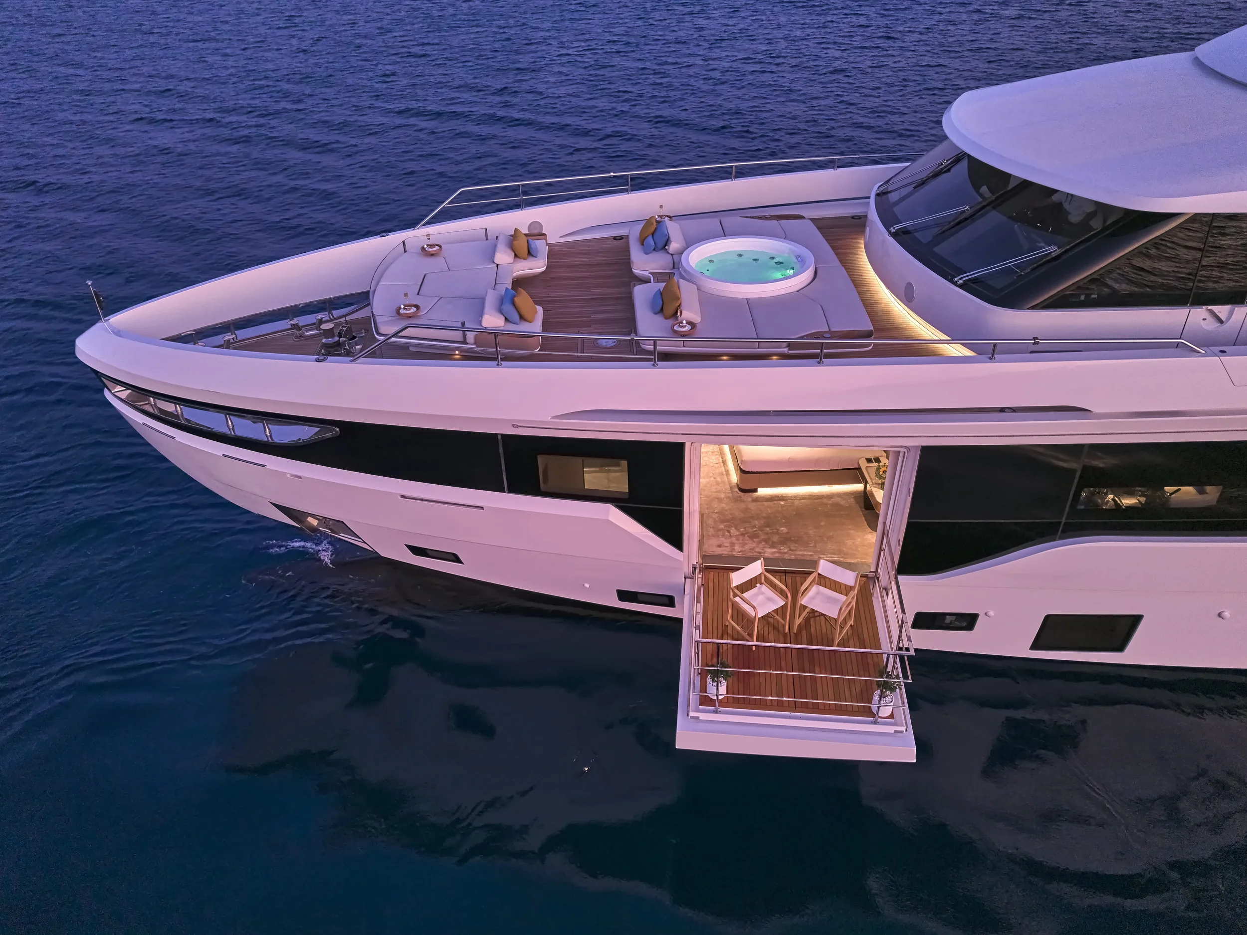 A luxury yacht sailing on the ocean with a deck area featuring seating, a hot tub, and a small outdoor dining area with two chairs.
