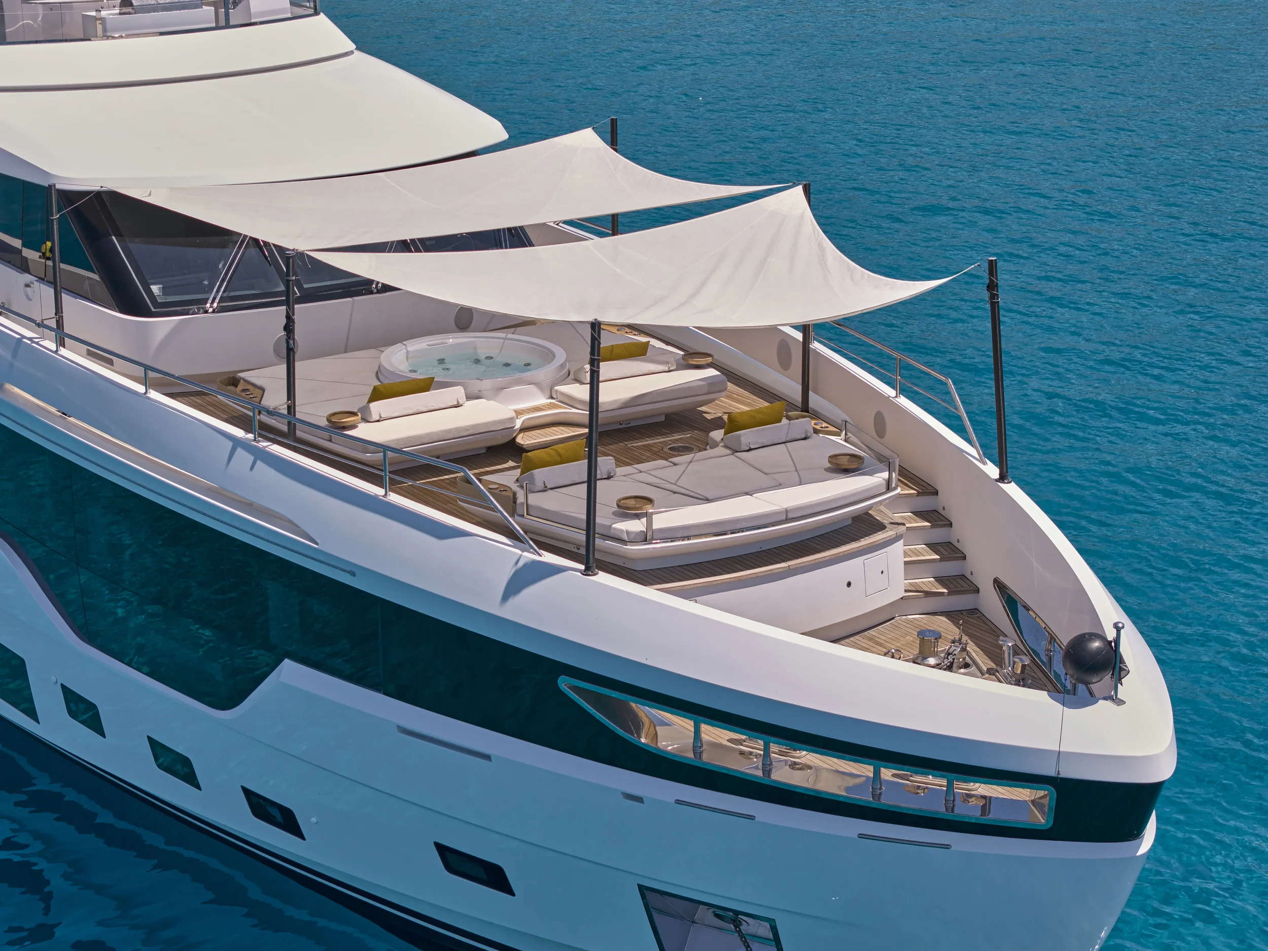 Luxury yacht with a deck area featuring sun loungers, a hot tub, and shade sails, floating on clear blue water.