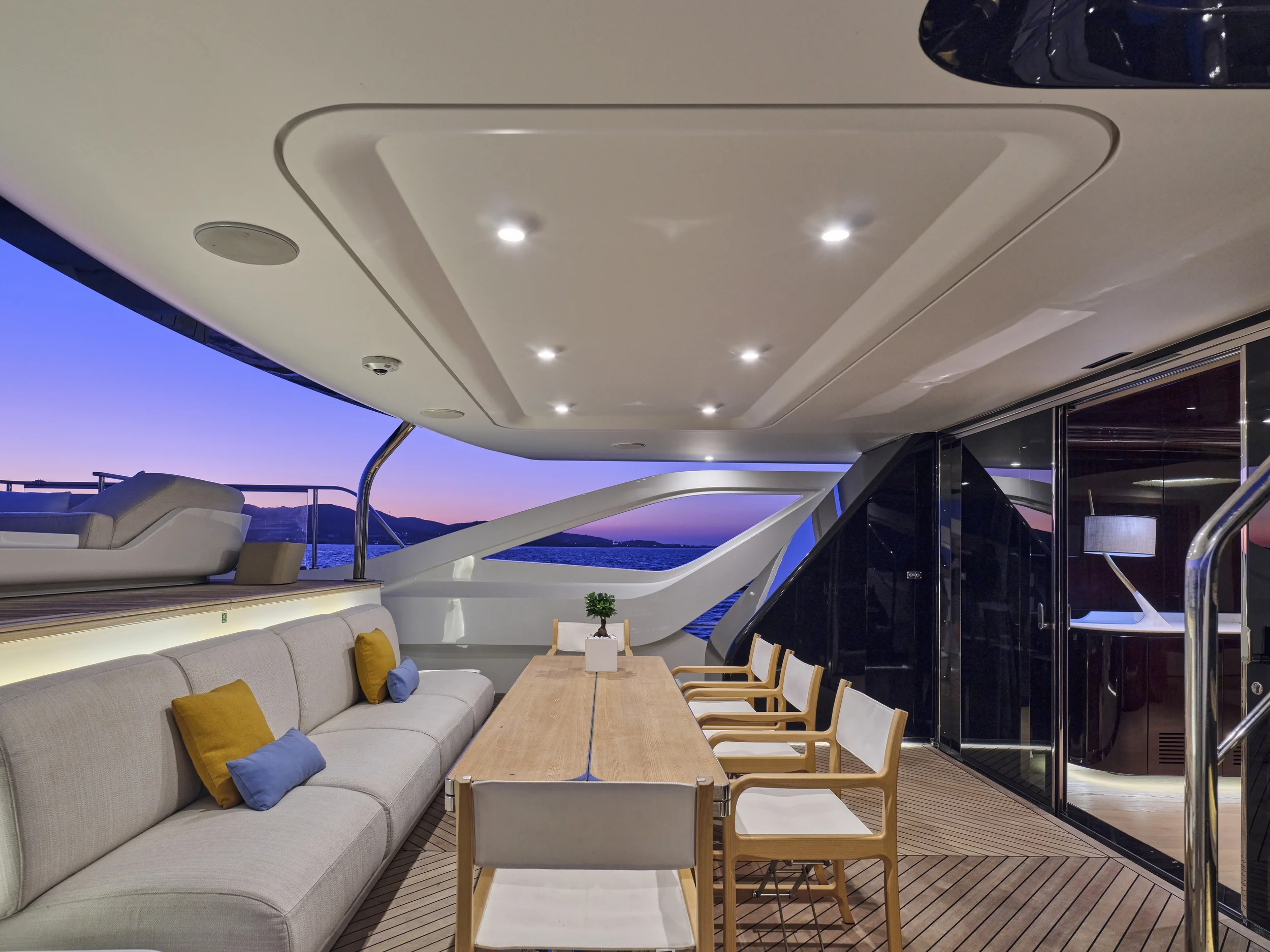 Luxurious yacht deck with outdoor dining area, beige sofa with colored pillows, wooden table with chairs, overlooking water and sunset.