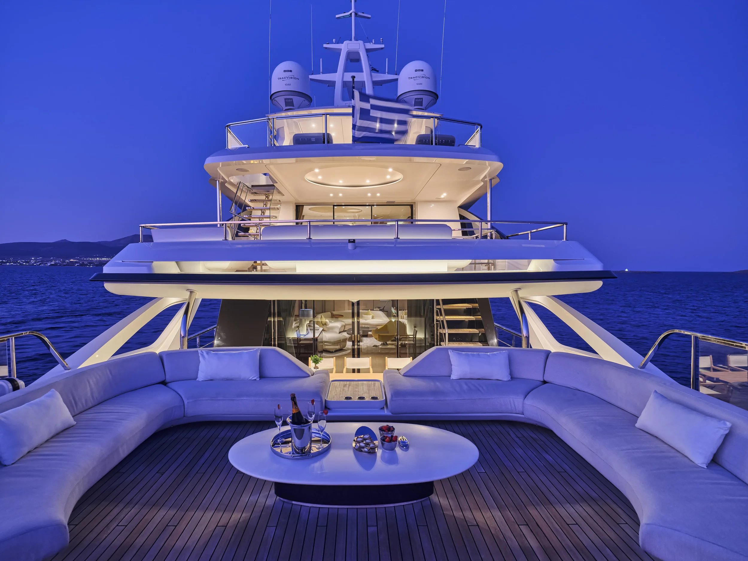 Luxury yacht at dusk with outdoor seating area, white curved couches, oval coffee table with drinks, and a large yacht structure in the background against a dark blue sky and water.