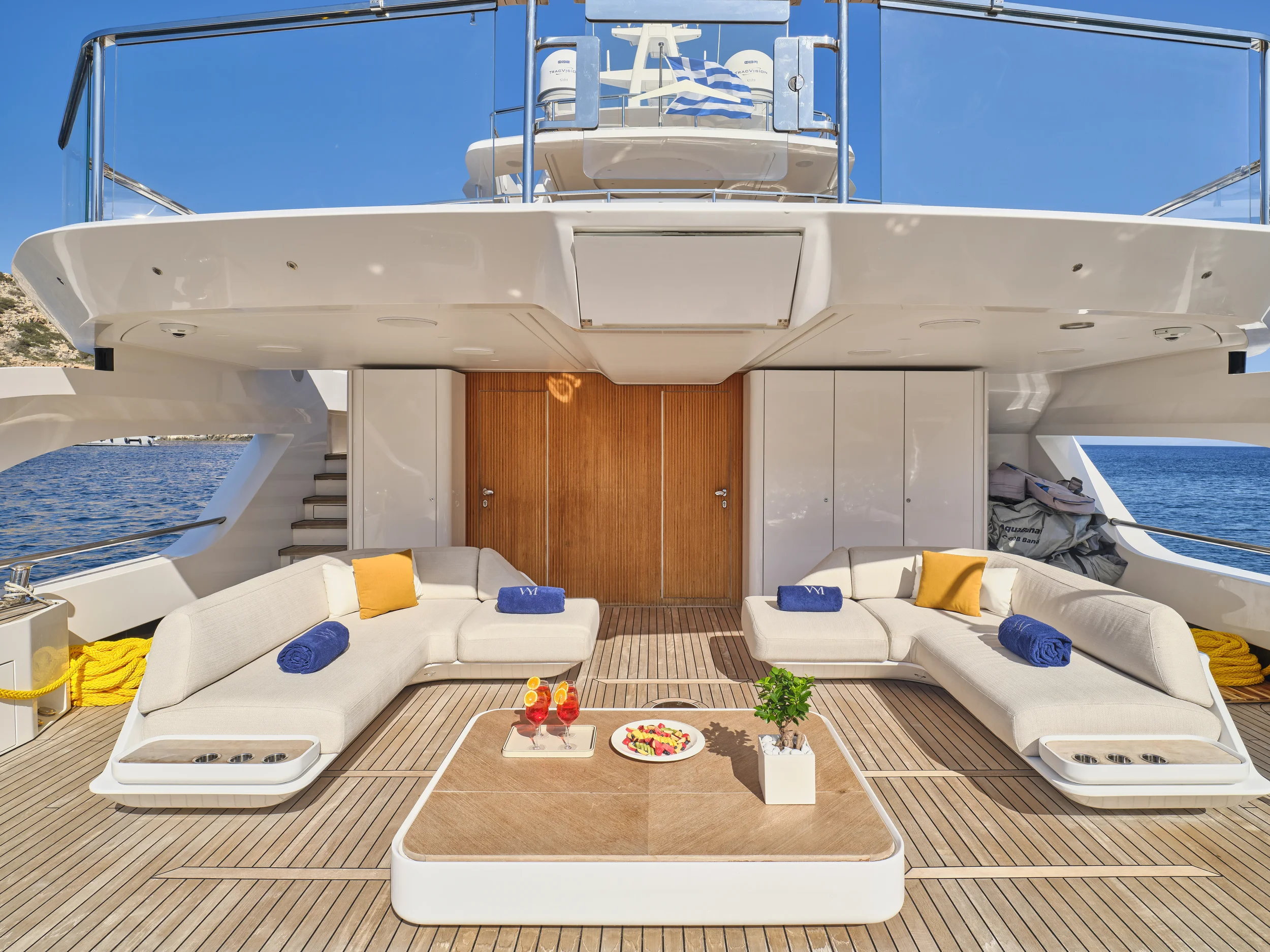 Luxury yacht deck with beige outdoor seating, blue and yellow towels, cocktails, a small plant, and clear blue sky with water in background.