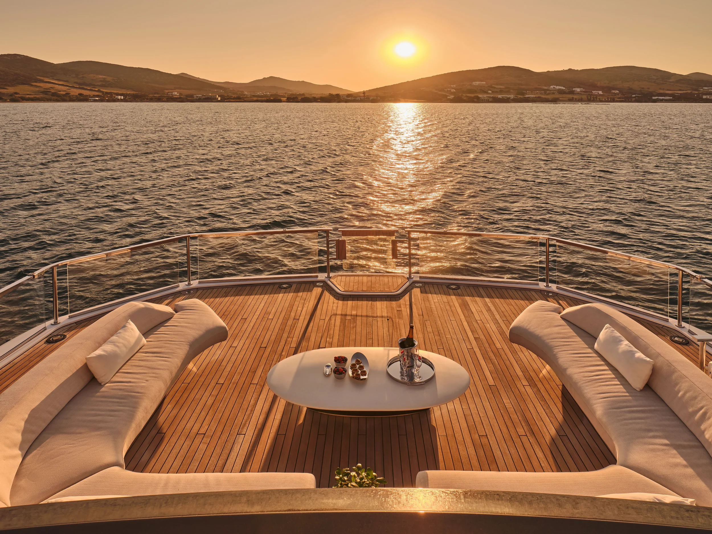 On the deck of a yacht during sunset, two beige curved lounge chairs with pillows face the water. A white coffee table in the center holds a bottle of champagne, glasses, and snacks. The ocean and distant hills are visible as the sun sets.