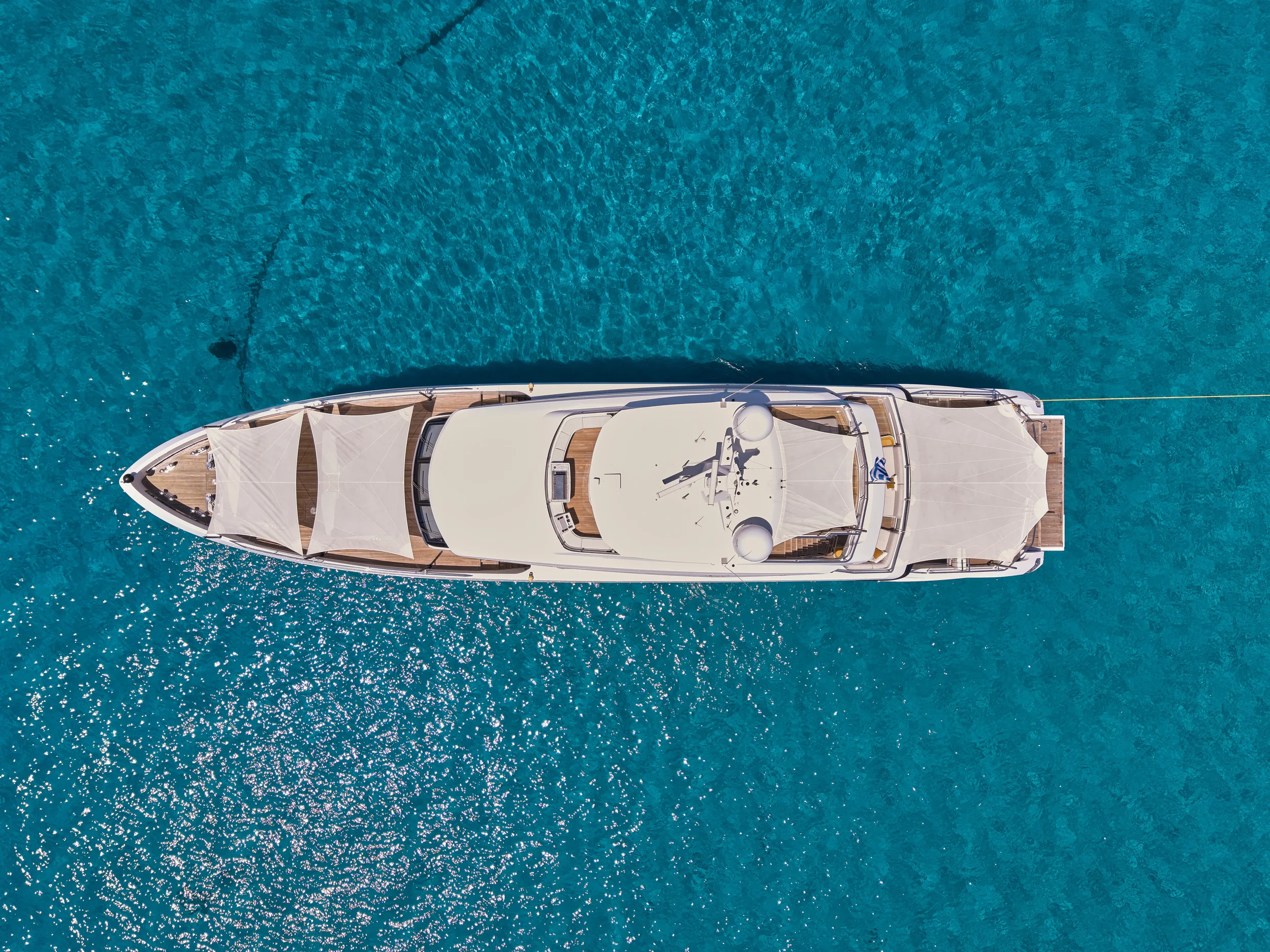 Aerial view of a luxury yacht floating on clear blue water, with a shaded area at the bow and a large deck at the stern.