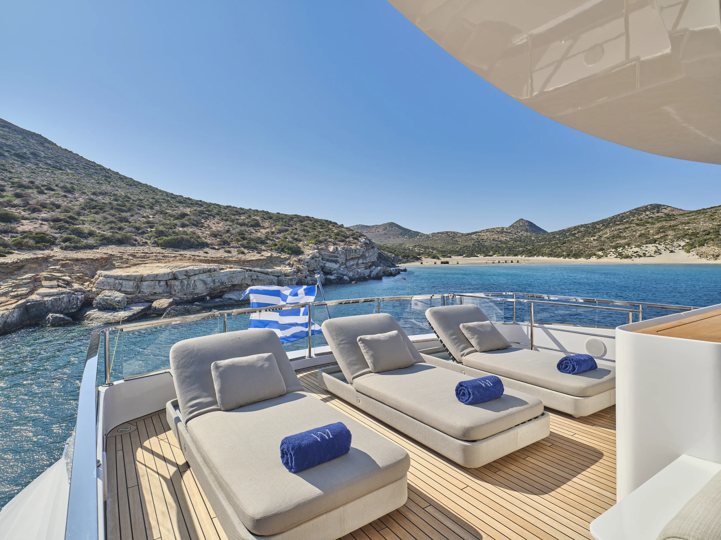 Sun loungers with rolled towels on a yacht deck overlooking a rocky coastline and blue water under a clear sky.