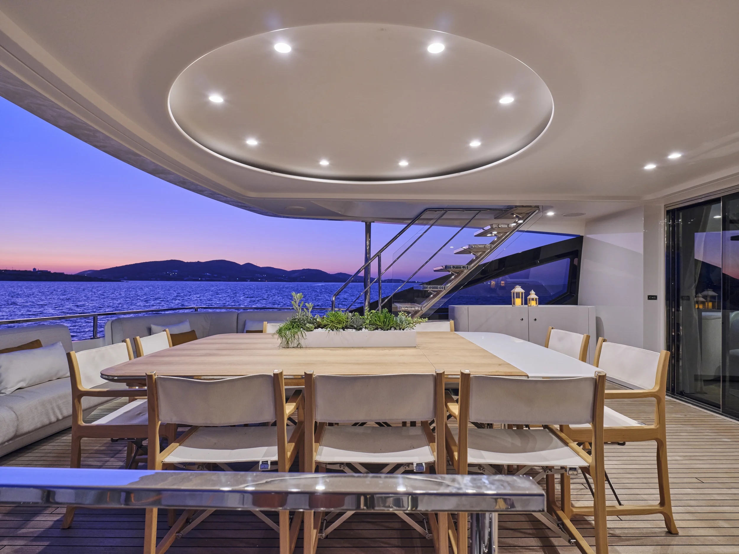 Empty dining area on a yacht with a large wooden table surrounded by beige and wooden chairs, overlooking a lake during sunset, with stairs leading up to an upper deck and decorative plants in the center of the table.