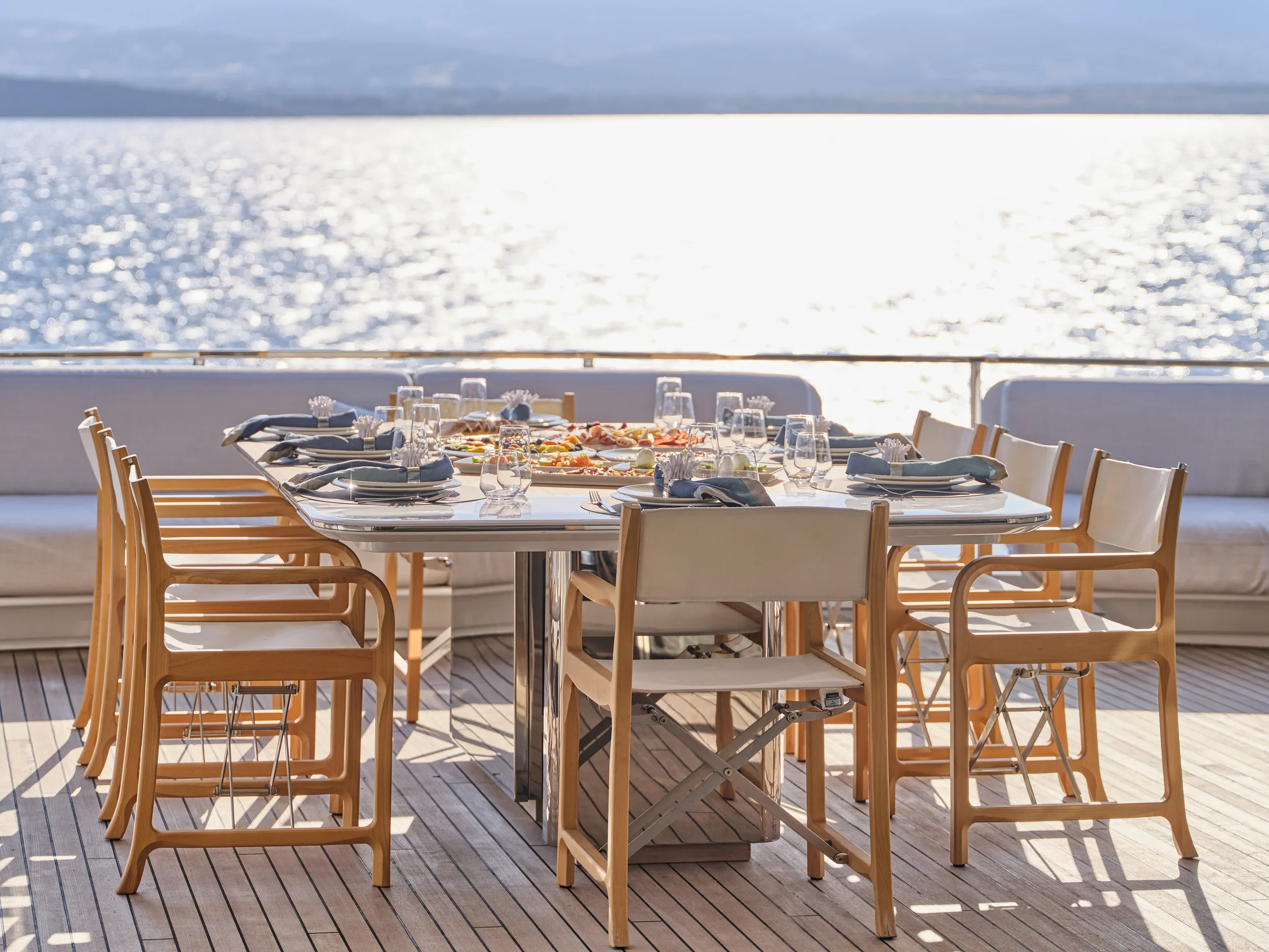 Dining table set with plates, napkins, glassware, and food on a boat deck near water.