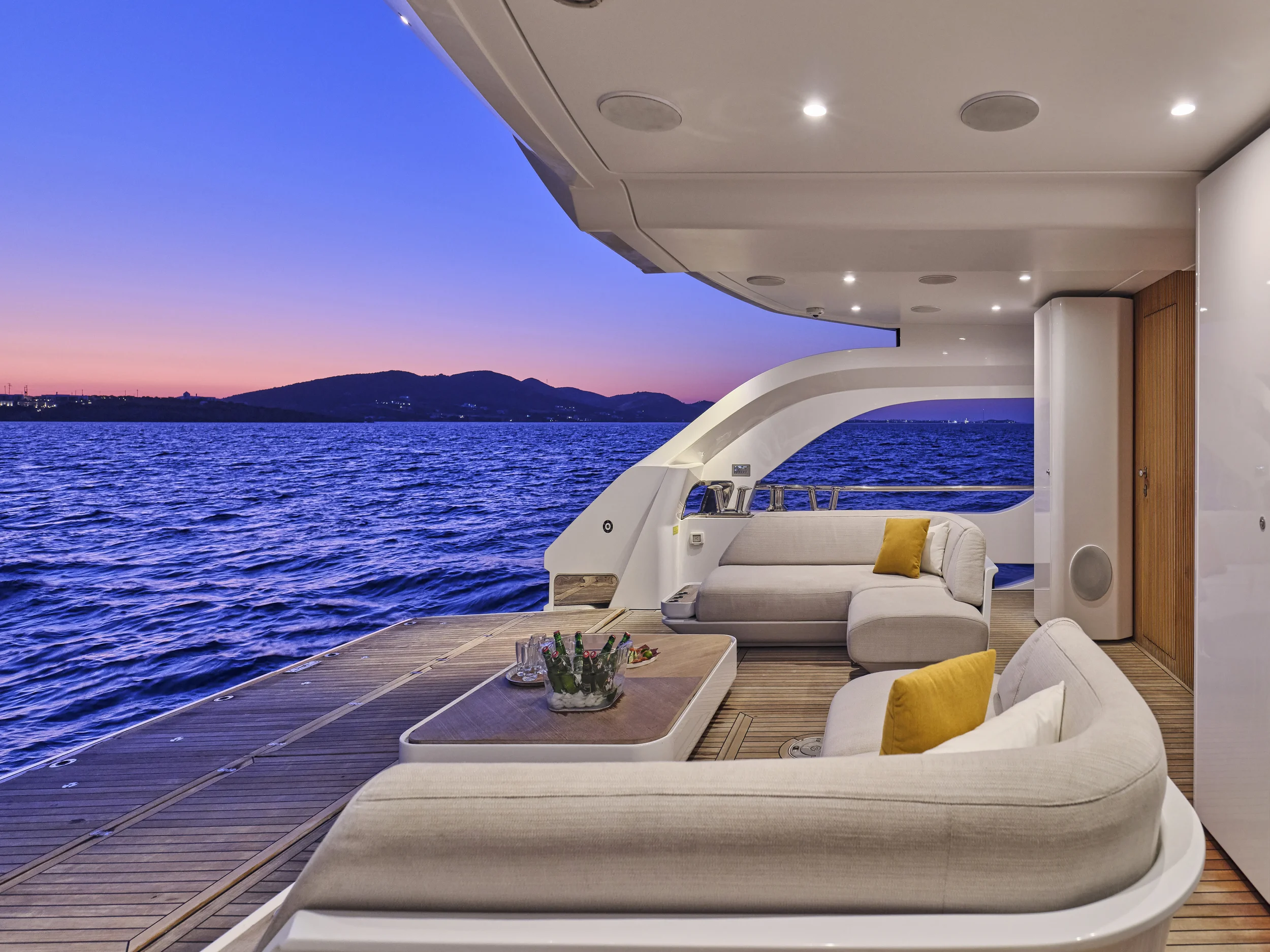 Luxury yacht balcony with beige outdoor sofas, yellow pillows, and a wooden table holding drinks and a bowl of snacks, overlooking calm blue water and distant hills during sunset.