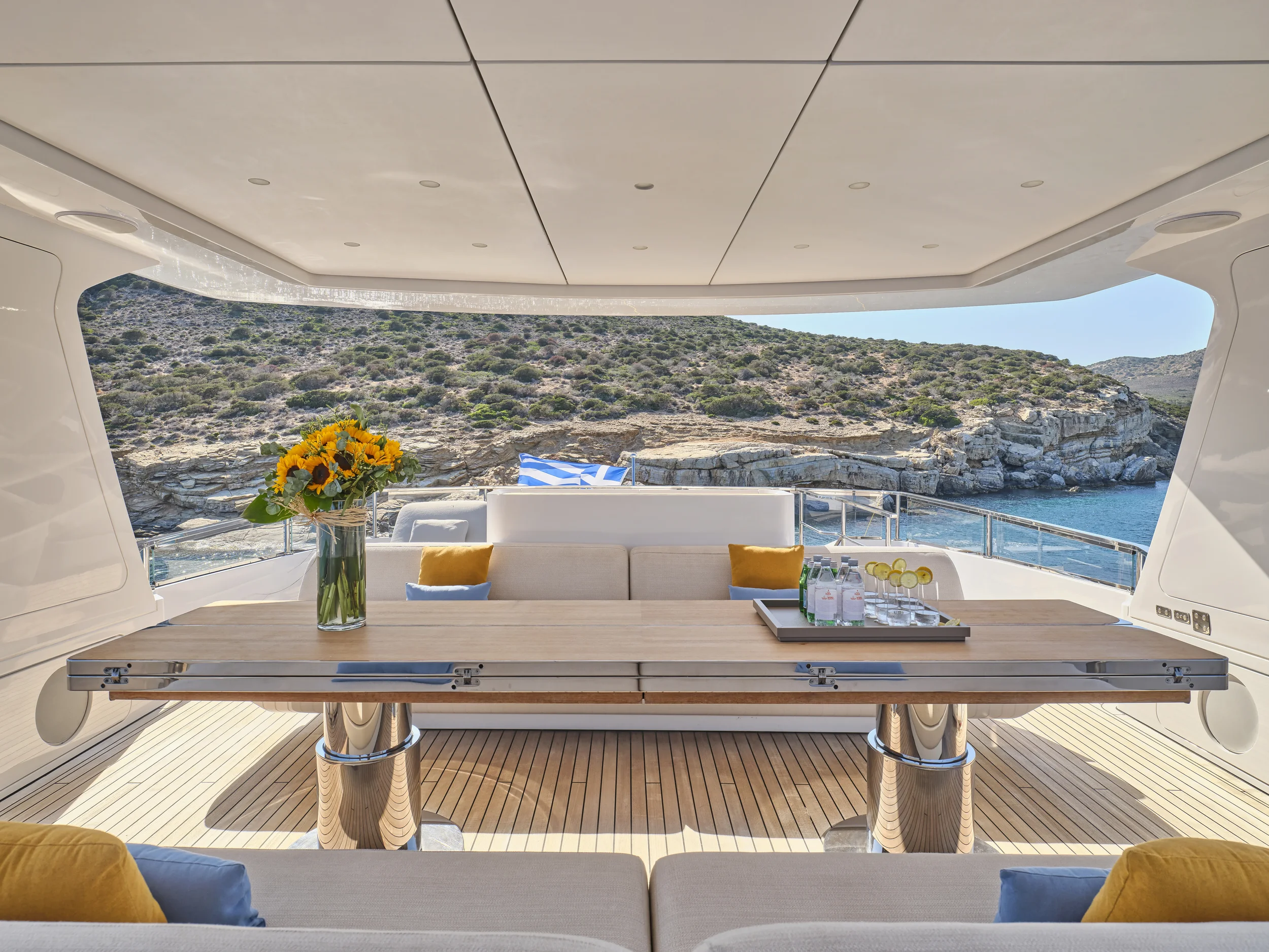 Luxury yacht deck with a wooden table, a vase with sunflowers, water bottles, and glasses, overlooking rocky shoreline and blue water.
