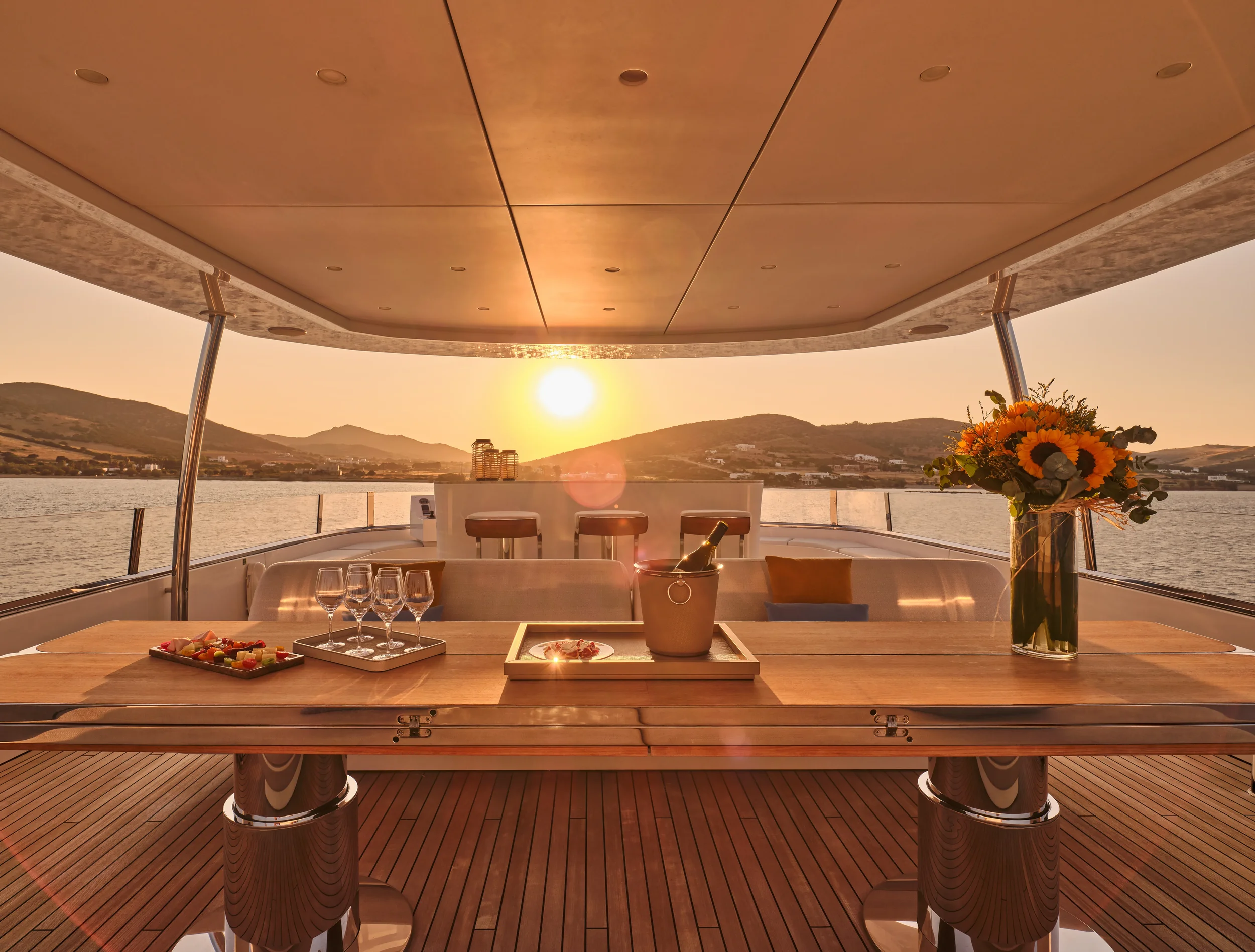 A luxurious yacht deck at sunset with a wooden table set with wine glasses, a tray of appetizers, an ice bucket with a bottle, and a vase of sunflowers, overlooking a calm body of water and distant hills.