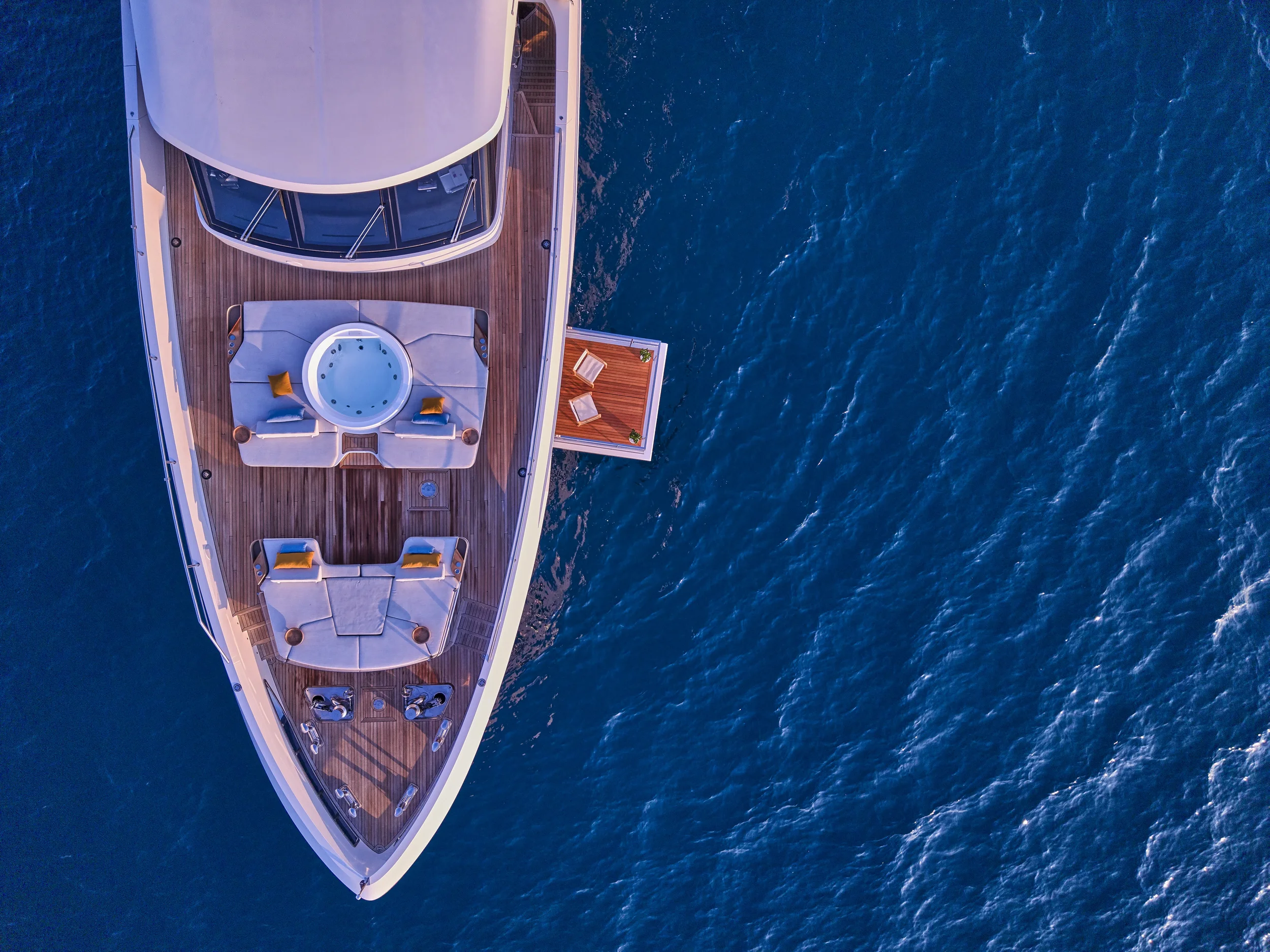 Aerial view of a luxury yacht's deck with seating area, hot tub, and lounge chairs, floating on blue ocean water.