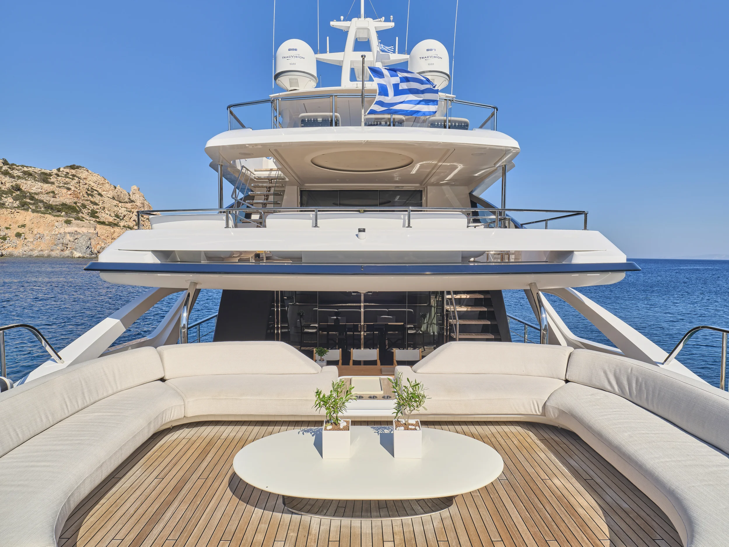 View from the deck of a luxury yacht showing a white curved outdoor seating area with a white table and two potted plants, with the sea and a rocky coastline in the background.