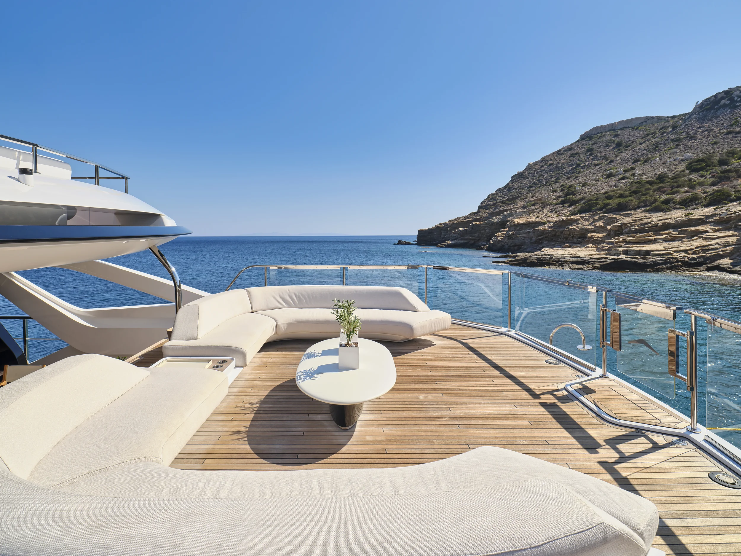 A yacht deck with white outdoor furniture, a small potted plant on a coffee table, overlooking a calm sea with rocky hills in the background under a clear blue sky.