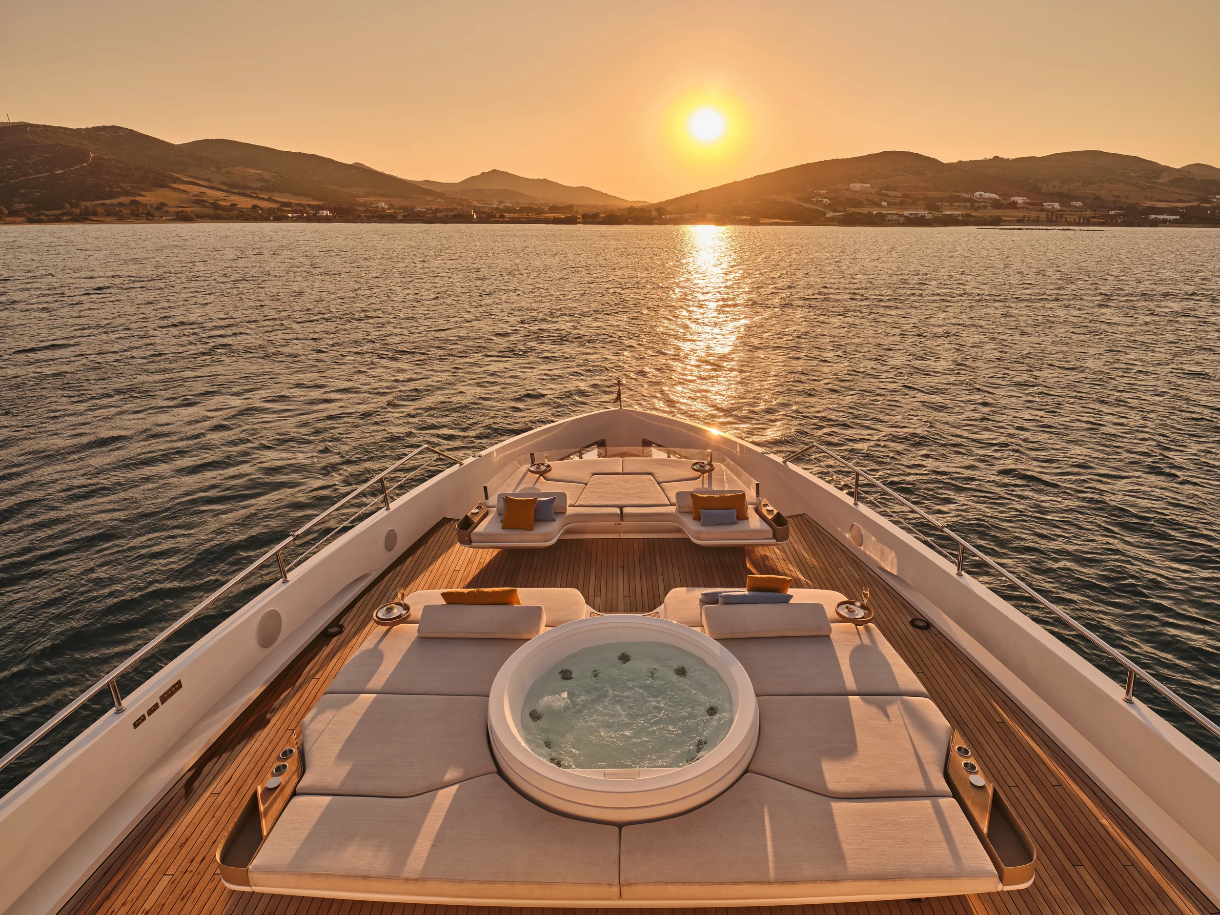A yacht with a hot tub and lounge chairs on the deck at sunset, overlooking a body of water and distant mountains.