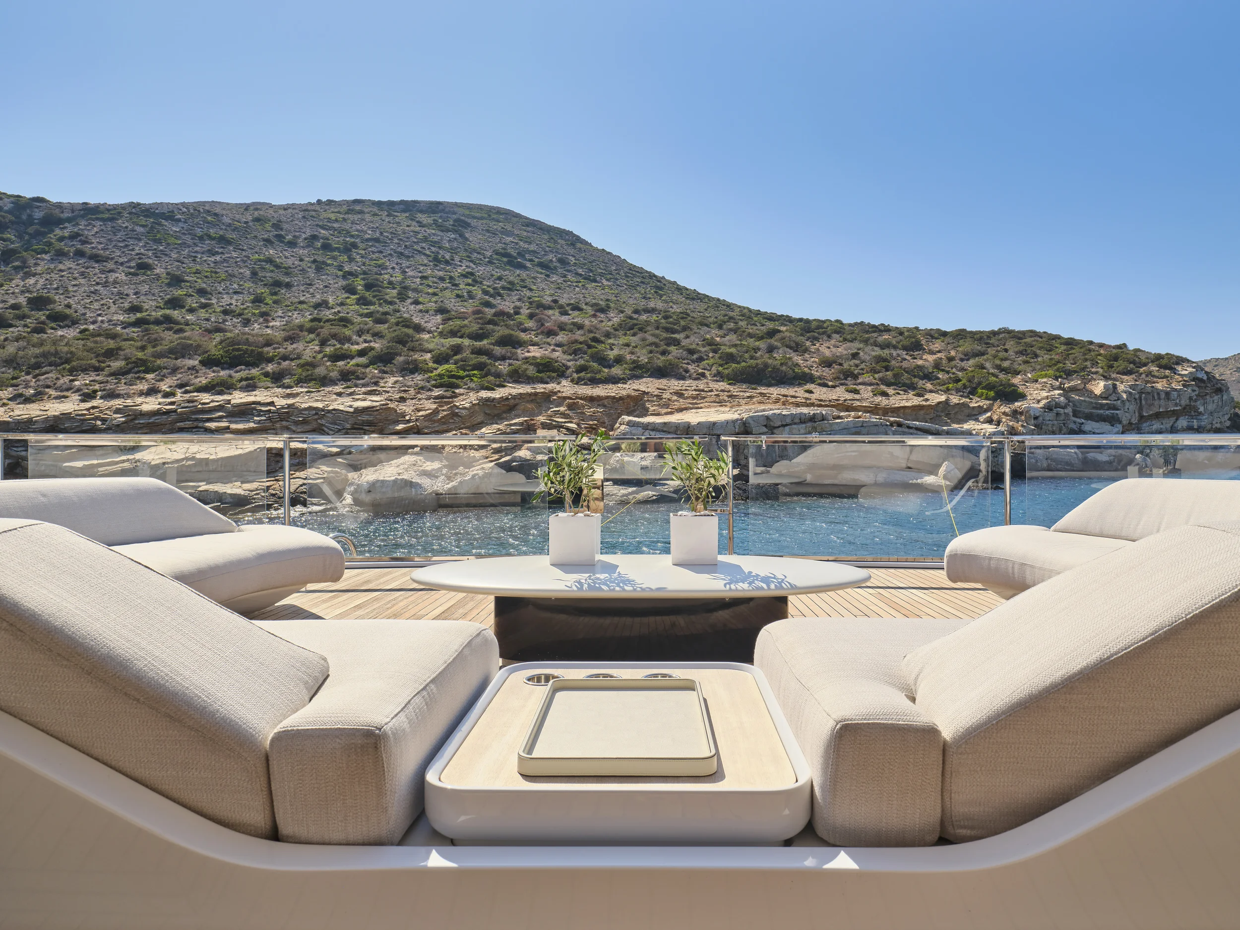 View of a boat deck with white cushioned seating, a round table with potted plants, overlooking rocky hillside and blue water under clear sky.