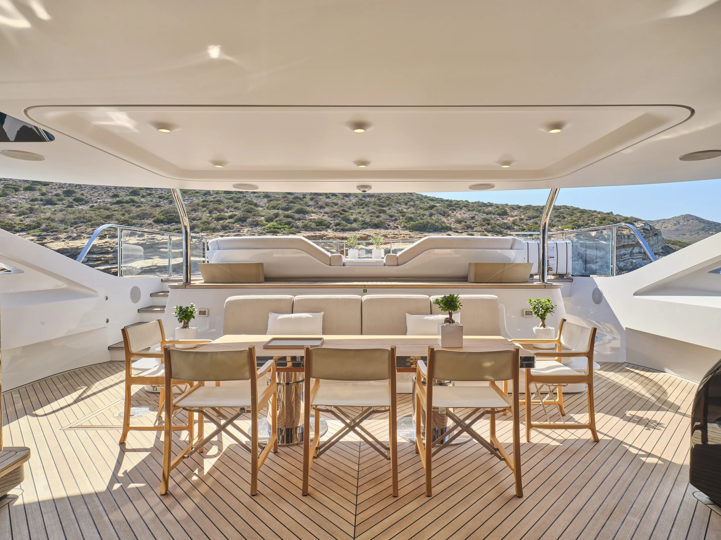 The upper deck of a yacht with beige seating, a dining table, and potted plants, overlooking a mountainous landscape.