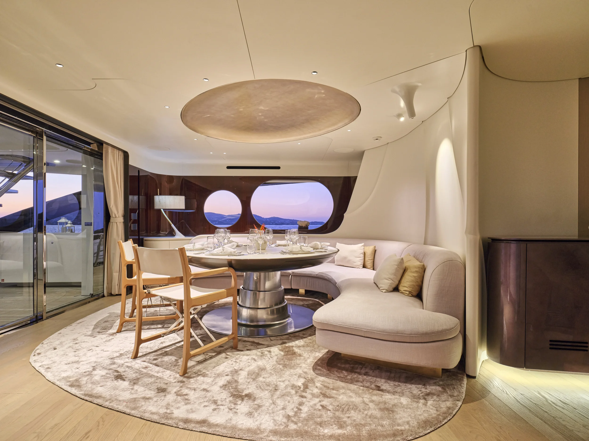 Luxurious yacht interior with a round dining table, white cushioned sofa, wooden chairs, and panoramic windows showing a sunset view.