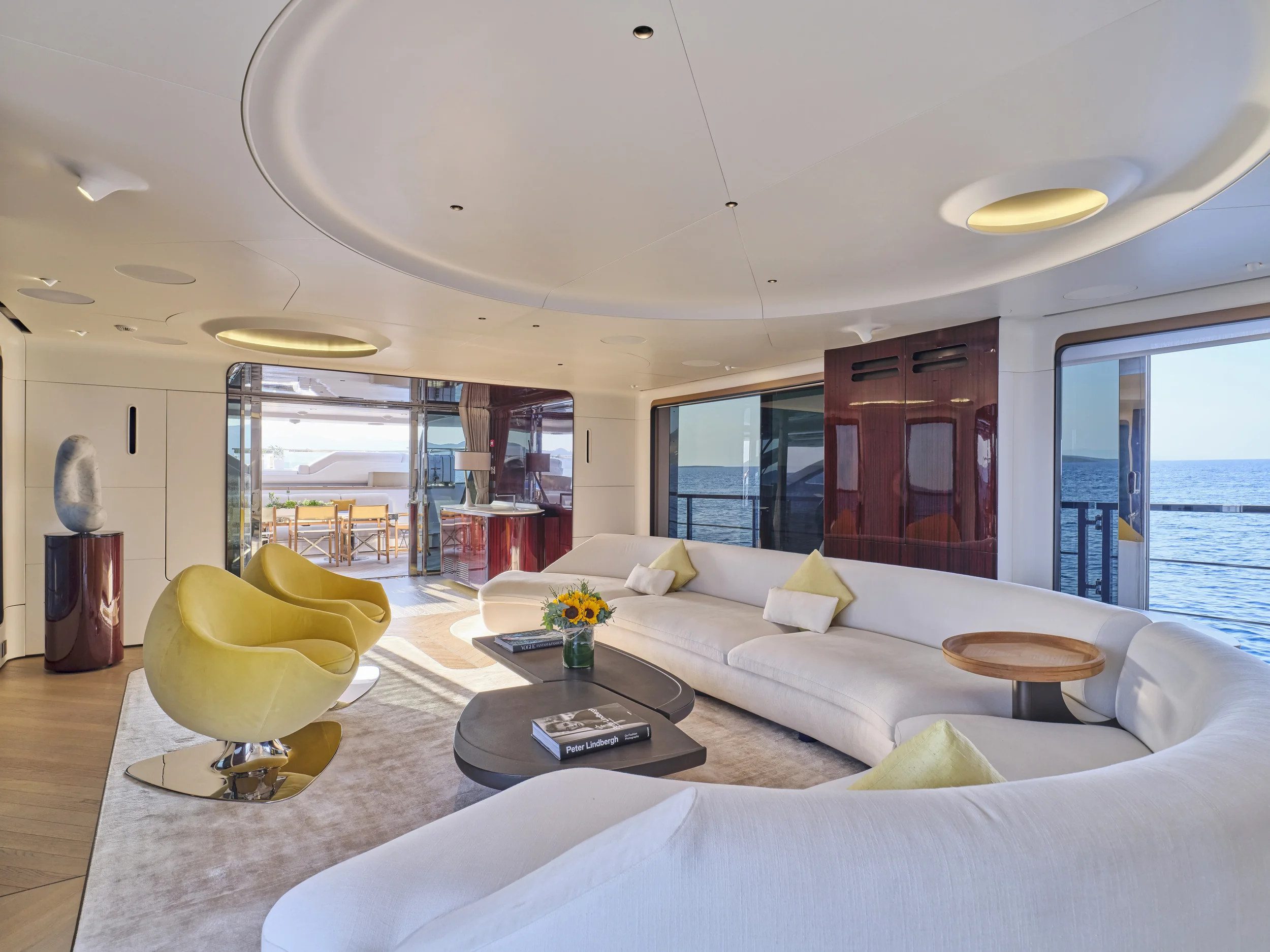 Luxury yacht interior living room with white sectional sofa, yellow chairs, coffee table with books and flowers, large windows showing ocean view, and balcony with outdoor seating.