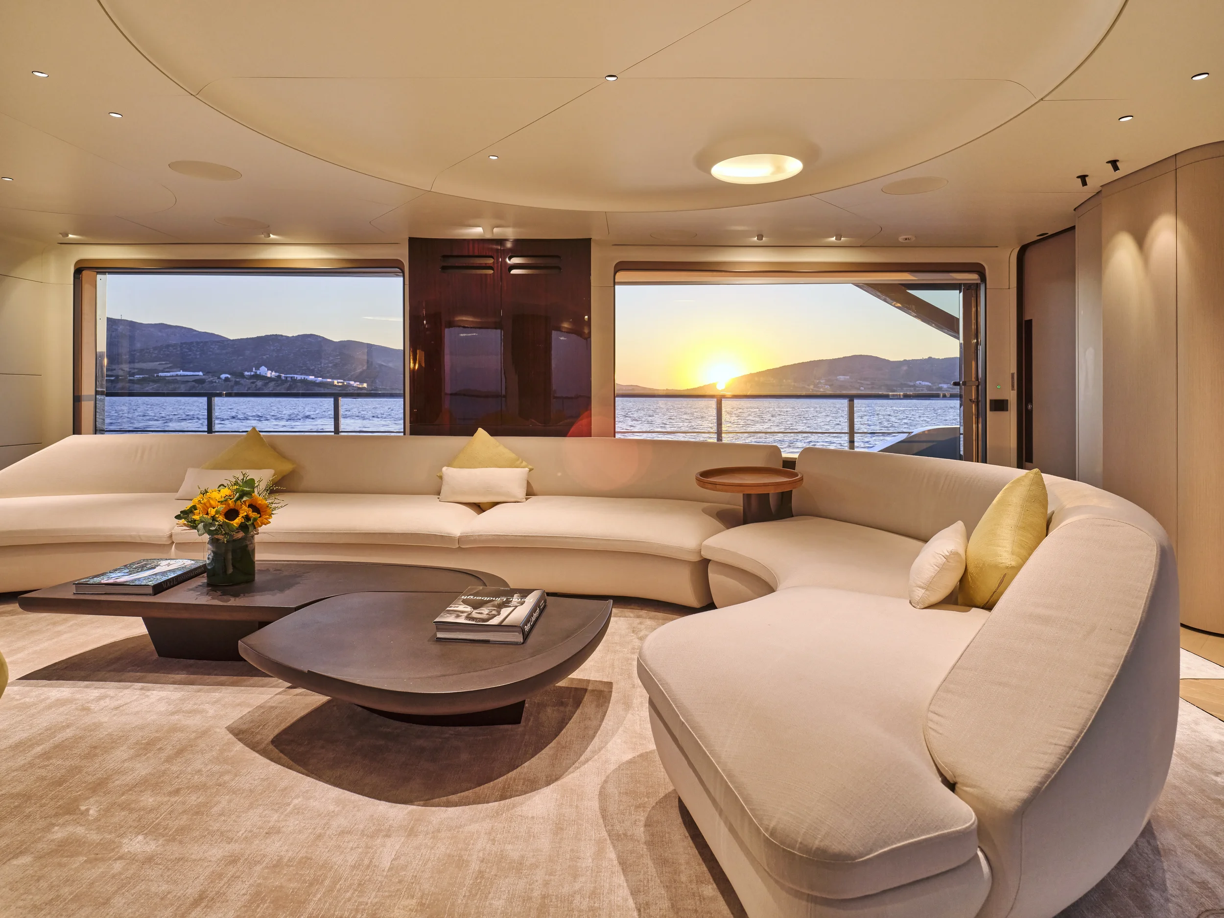 A luxurious living room with large windows overlooking a body of water and mountains at sunset, featuring beige curved sofas, a dark wood coffee table with books and a vase of sunflowers, a round wooden side table, and soft lighting.