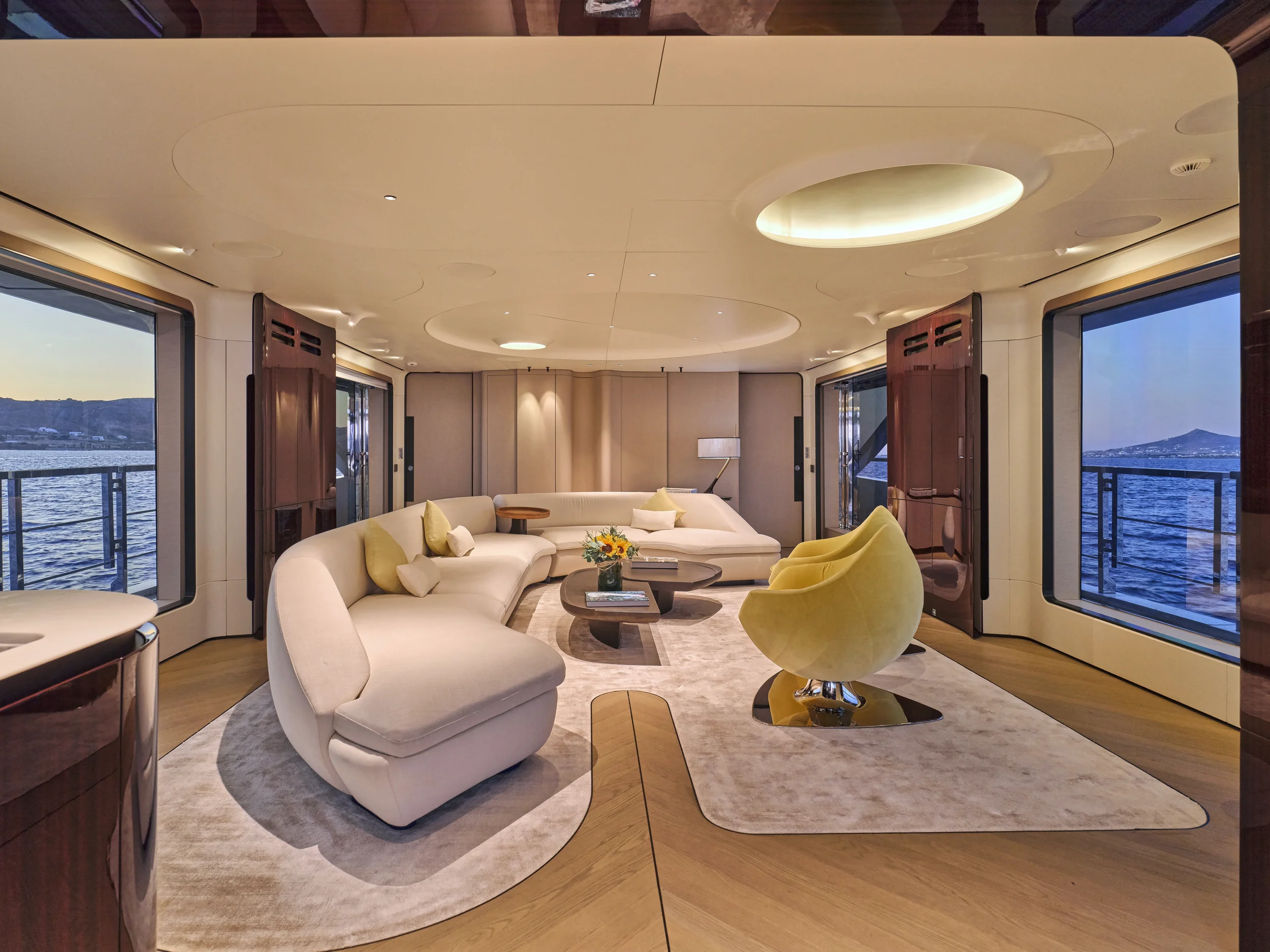 Luxurious living room on a yacht with large windows overlooking the water, featuring beige and yellow furniture, a round coffee table with a flower arrangement, and modern lighting fixtures.