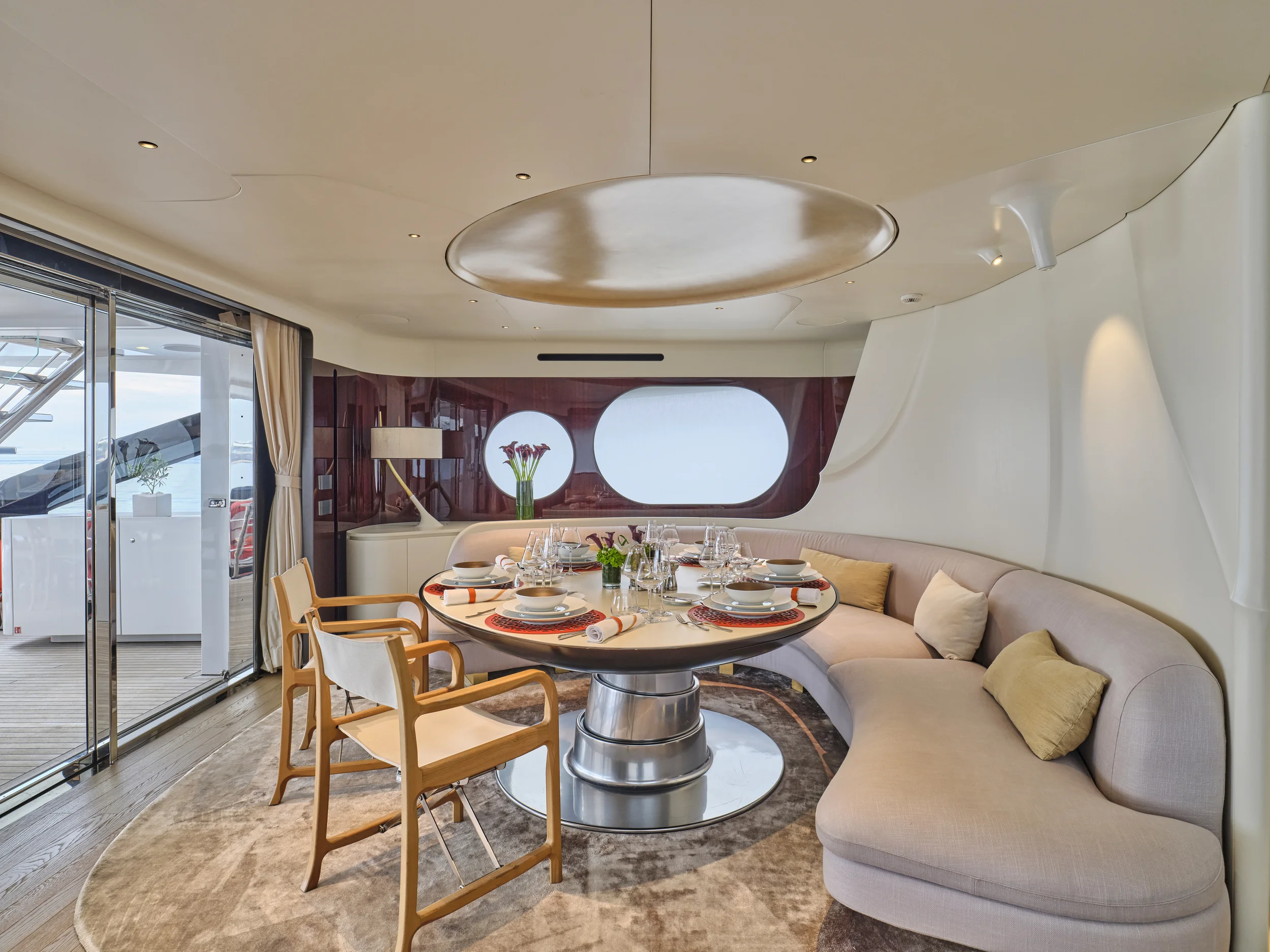 Elegant dining room on a yacht with a round table set for dinner, surrounded by beige seating and wooden chairs, with large windows and sliding glass door showing outdoor deck and ocean view.