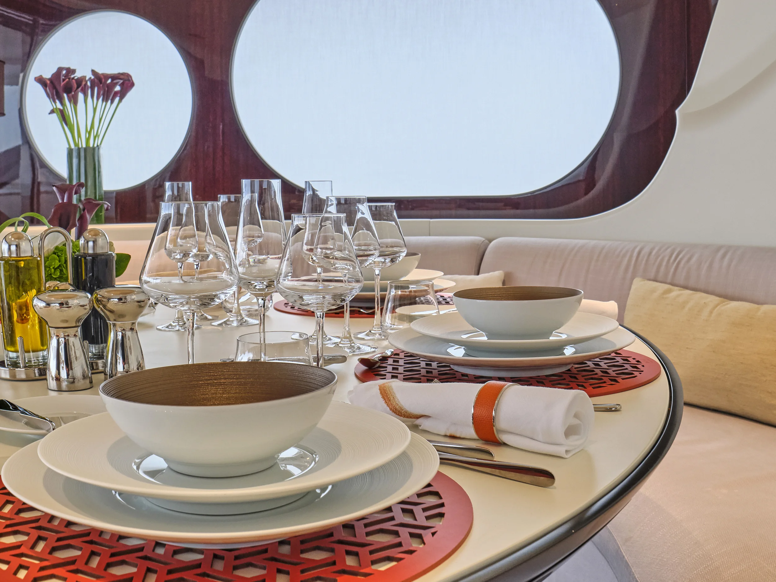 A finely set dining table with white plates, bowls, and various glasses, including wine glasses, on red placemats. There are silver salt and pepper shakers, oil and vinegar bottles, and a rolled-up napkin with a leather band. In the background, a win