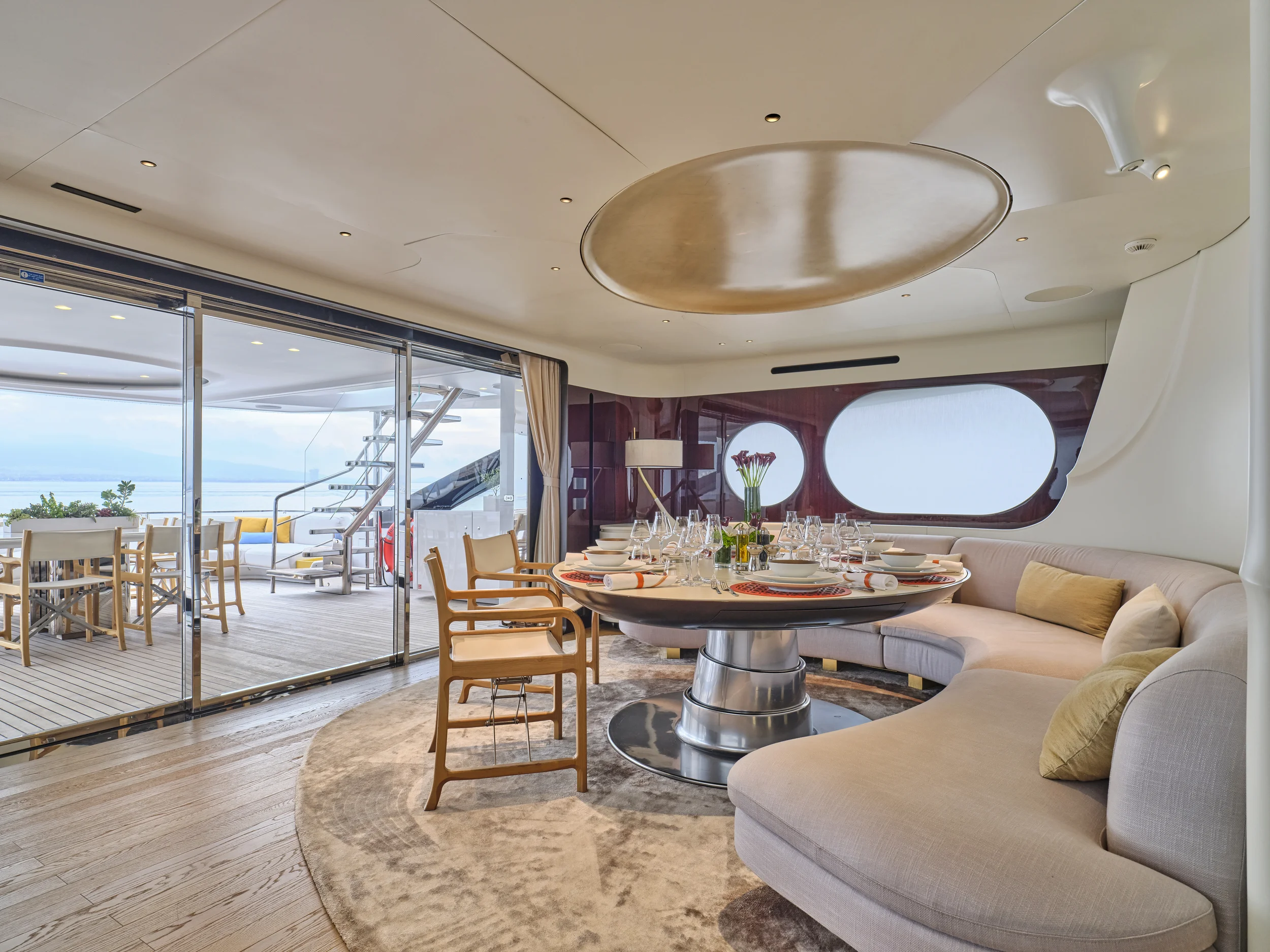 Elegant dining area on a yacht with a round table set for a meal, surrounded by a curved beige sofa, with large windows and a sliding glass door leading to a balcony with outdoor seating and a view of the water.