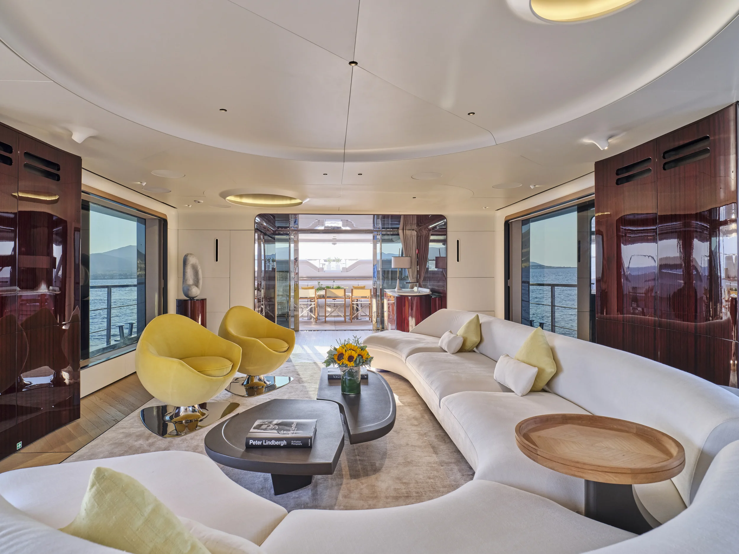 Luxurious living room with curved white sofa, yellow lounge chairs, a coffee table with a book and a vase of sunflowers, large windows overlooking the water, and a balcony in the background.