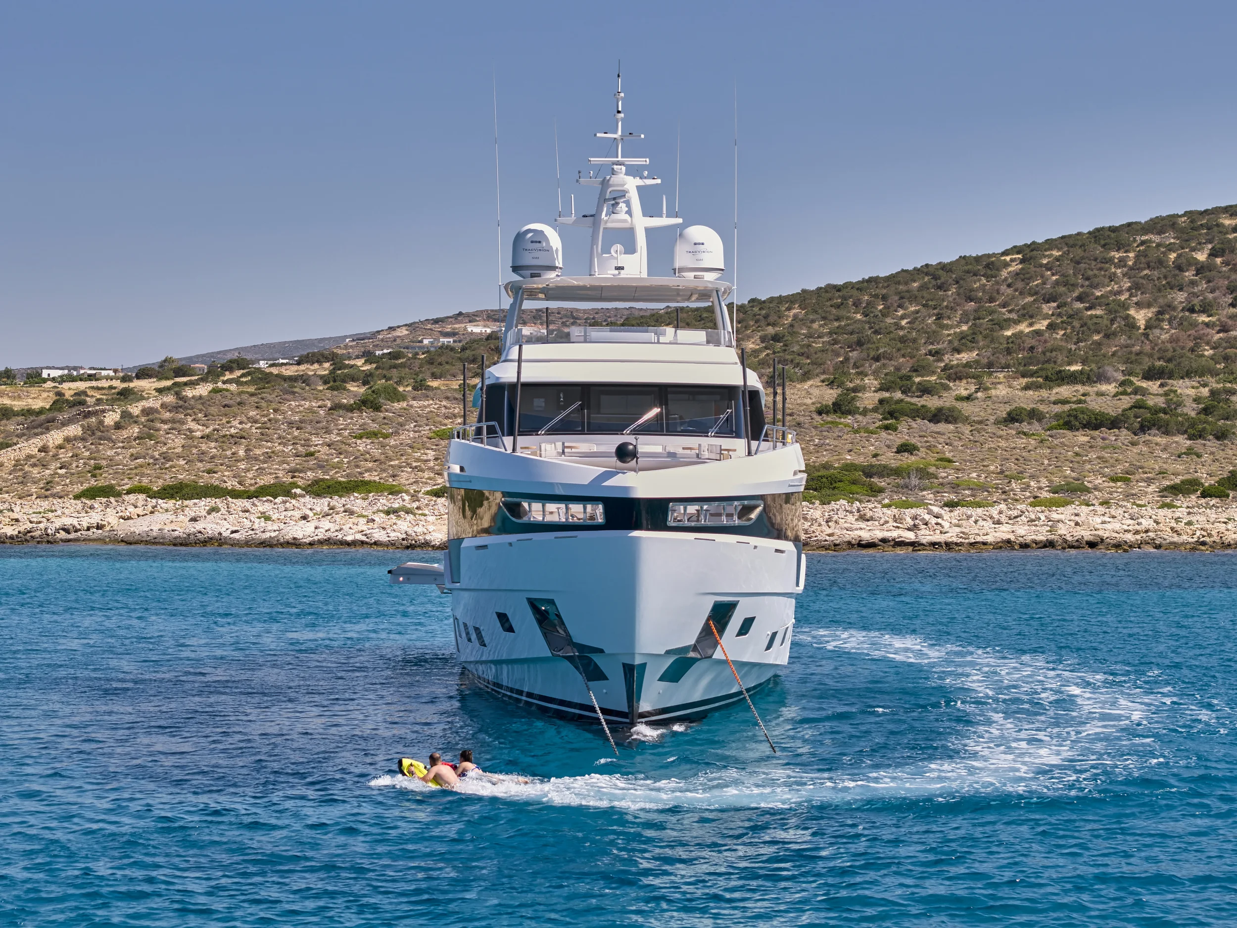 A large white yacht on the water, with two people swimming nearby, and a hilly, rocky coastline in the background.
