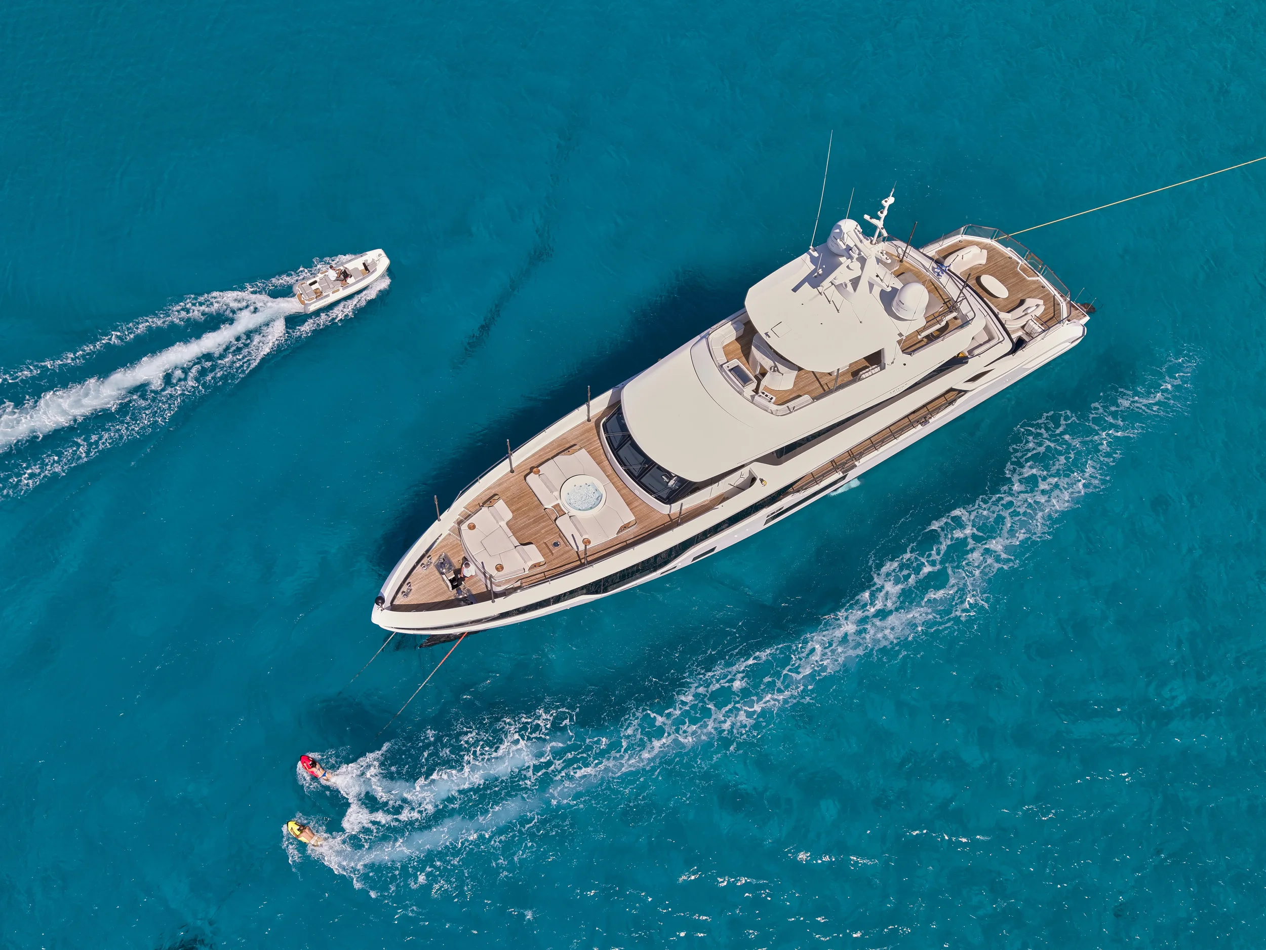 Aerial view of a large white yacht with brown decks sailing in bright blue water, accompanied by a small speedboat and two people on jet skis.