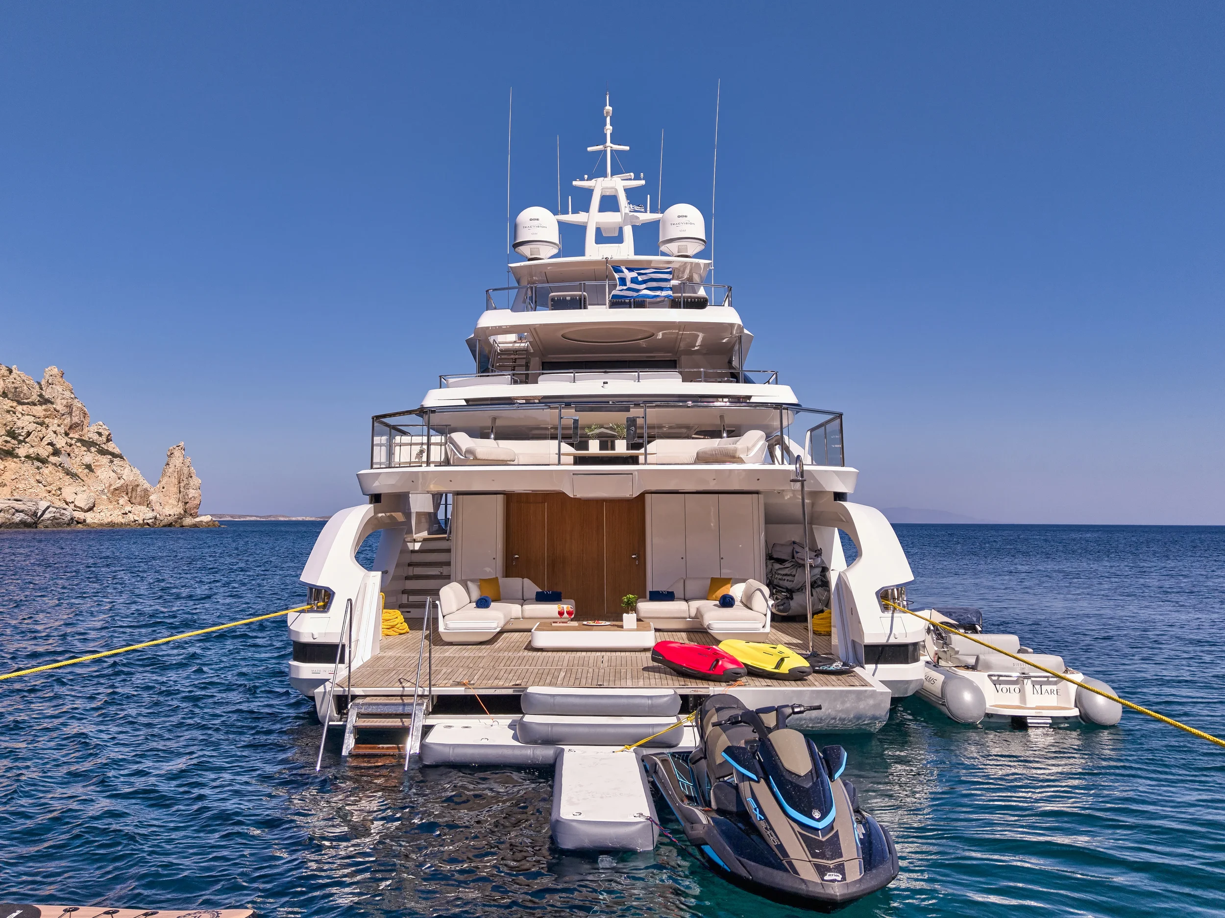 A luxury yacht docked at a pier on the water with a rocky coastline visible in the background. The yacht has multiple decks, outdoor seating, and watercraft including jet skis and small boats on the stern. The sky is clear and blue.