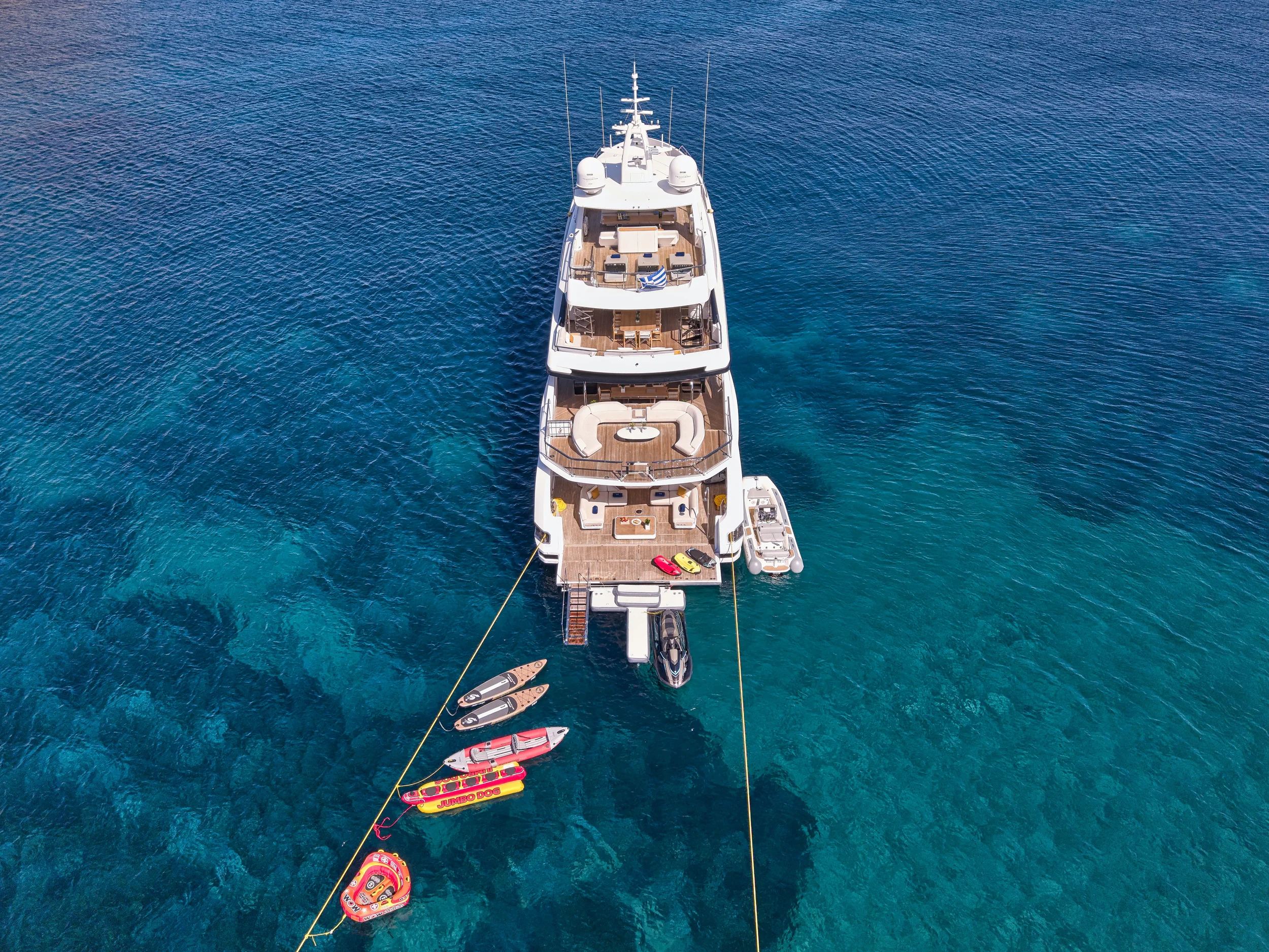 Aerial view of a large white yacht anchored in clear blue water with two smaller boats and inflatable water toys nearby.