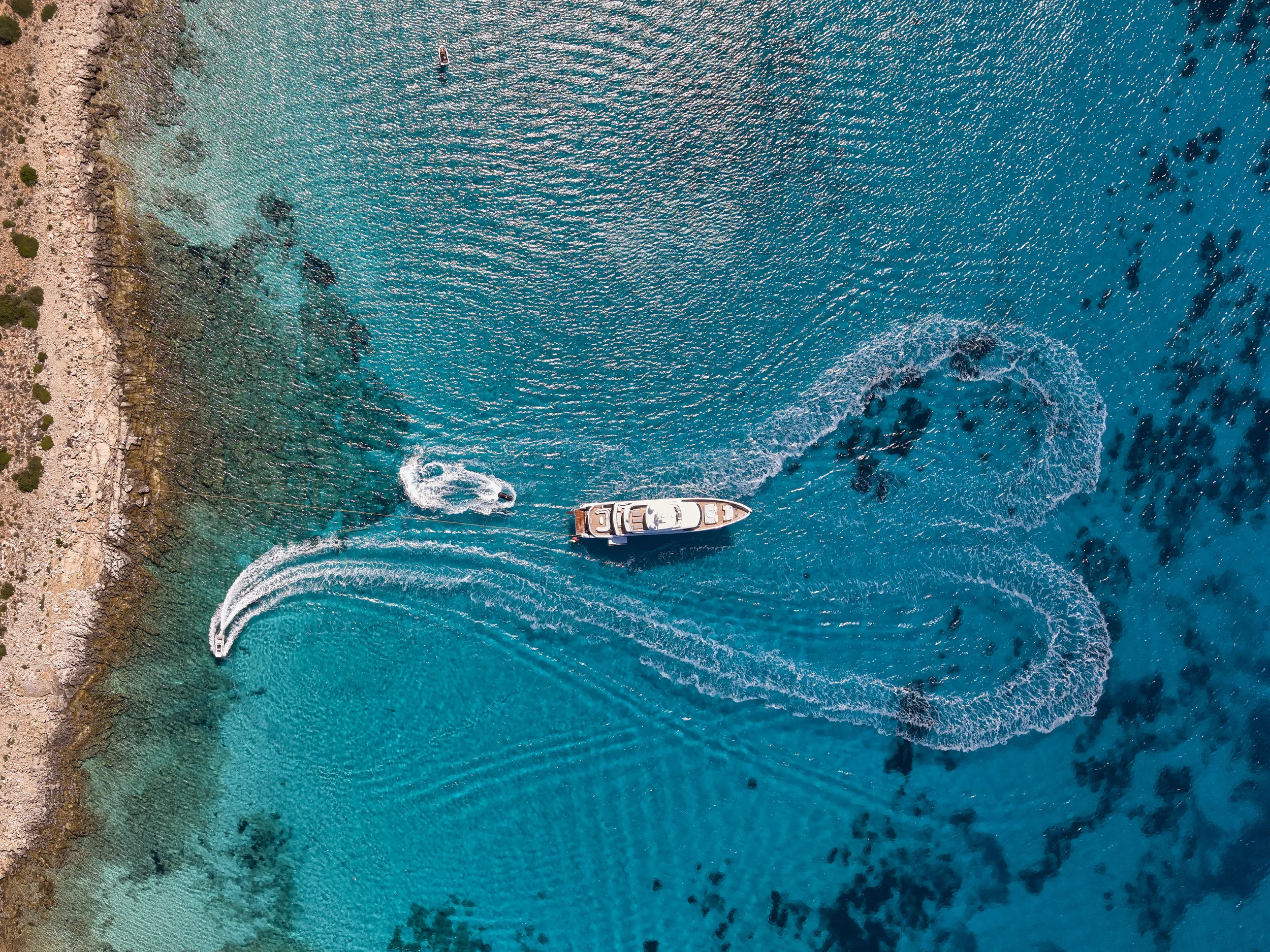 An aerial view of a boat and smaller watercraft on clear blue water near a rocky shoreline.