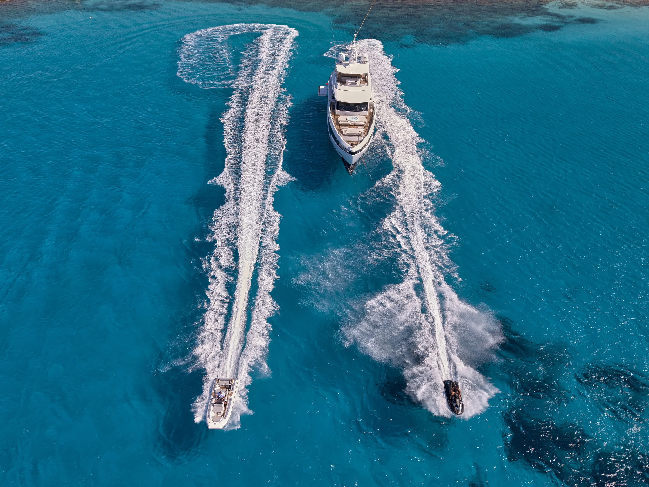 An aerial view of a large white yacht in the center with two smaller boats on either side, all moving in the same direction across bright blue water, creating wakes behind them