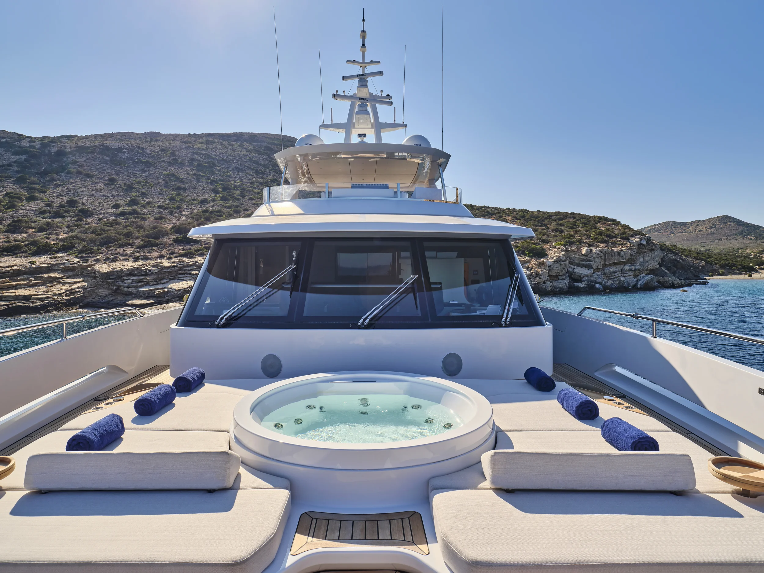 View of the front deck of a luxury yacht with built-in hot tub, sun loungers with rolled towels, in a scenic coastal area with rocks and hills in the background, under clear blue skies.