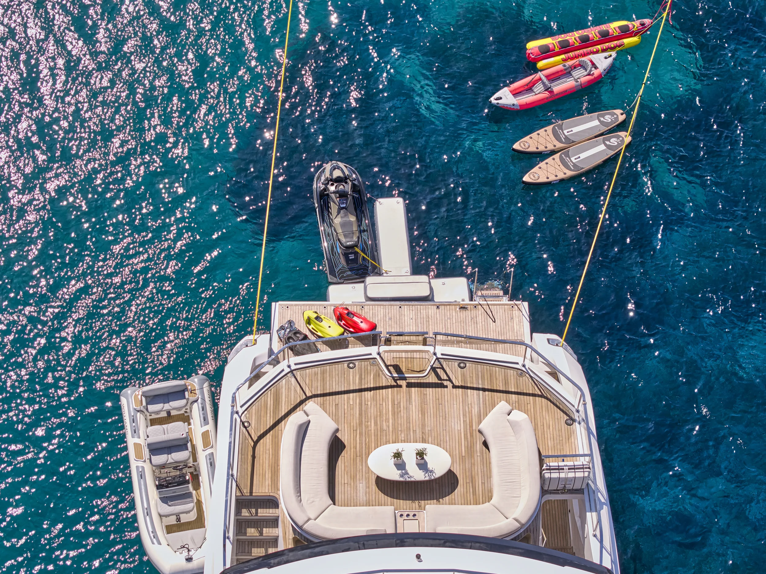 An aerial view of a yacht on the water with jet skis and inflatable water slides. The yacht has a wooden deck with seating, a table, and watercraft docked nearby.
