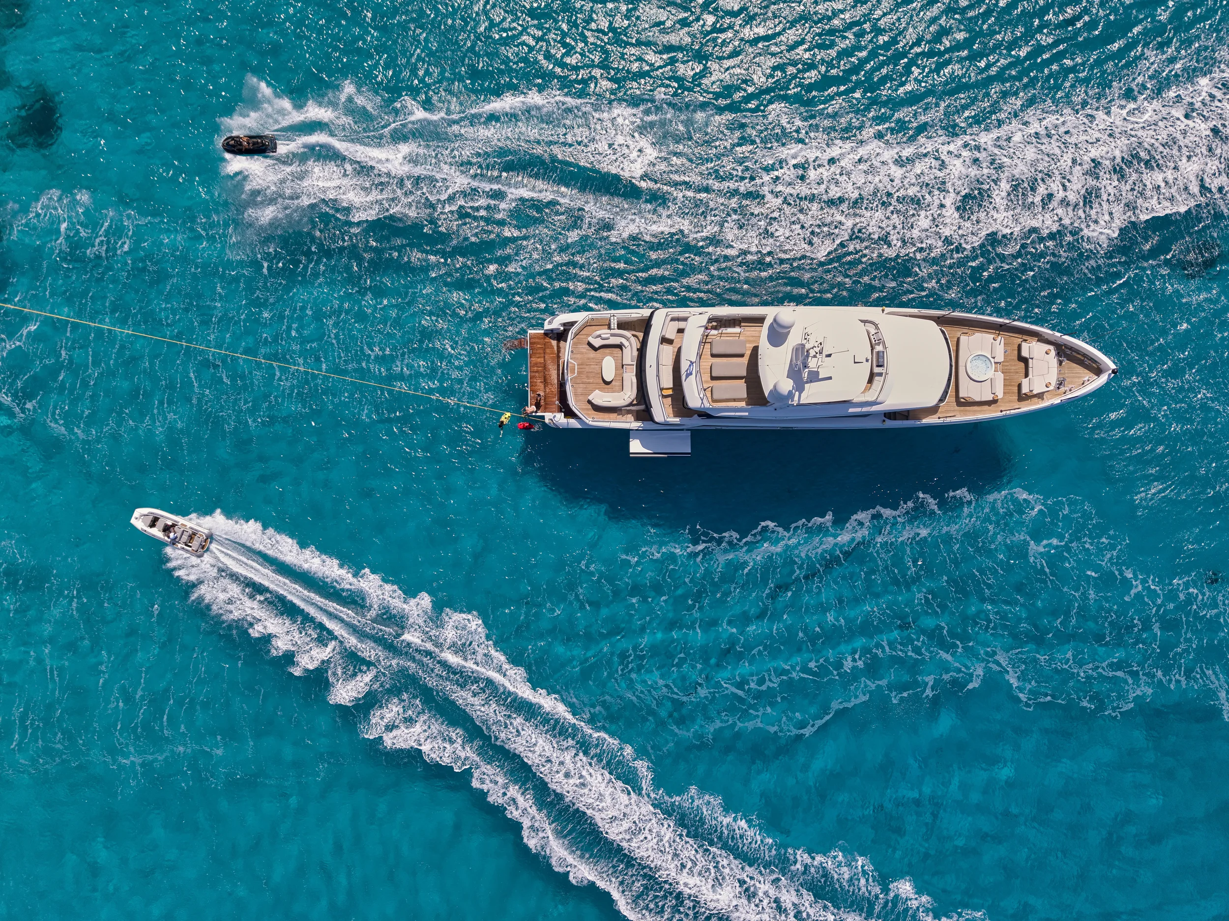 An aerial view of a large white yacht moving through clear blue water, with two smaller boats nearby.