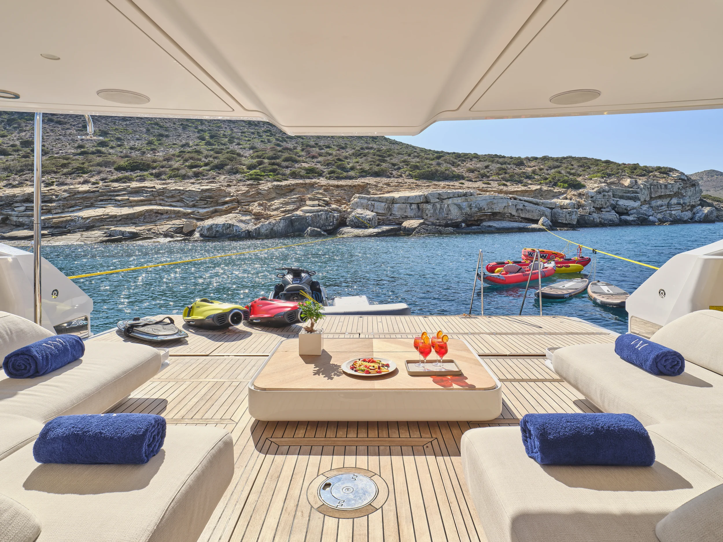View from a yacht deck overlooking blue water, rocky coastline, and a small raft with inflatable boats and jet skis, with shaded seating area and towels on lounge chairs.