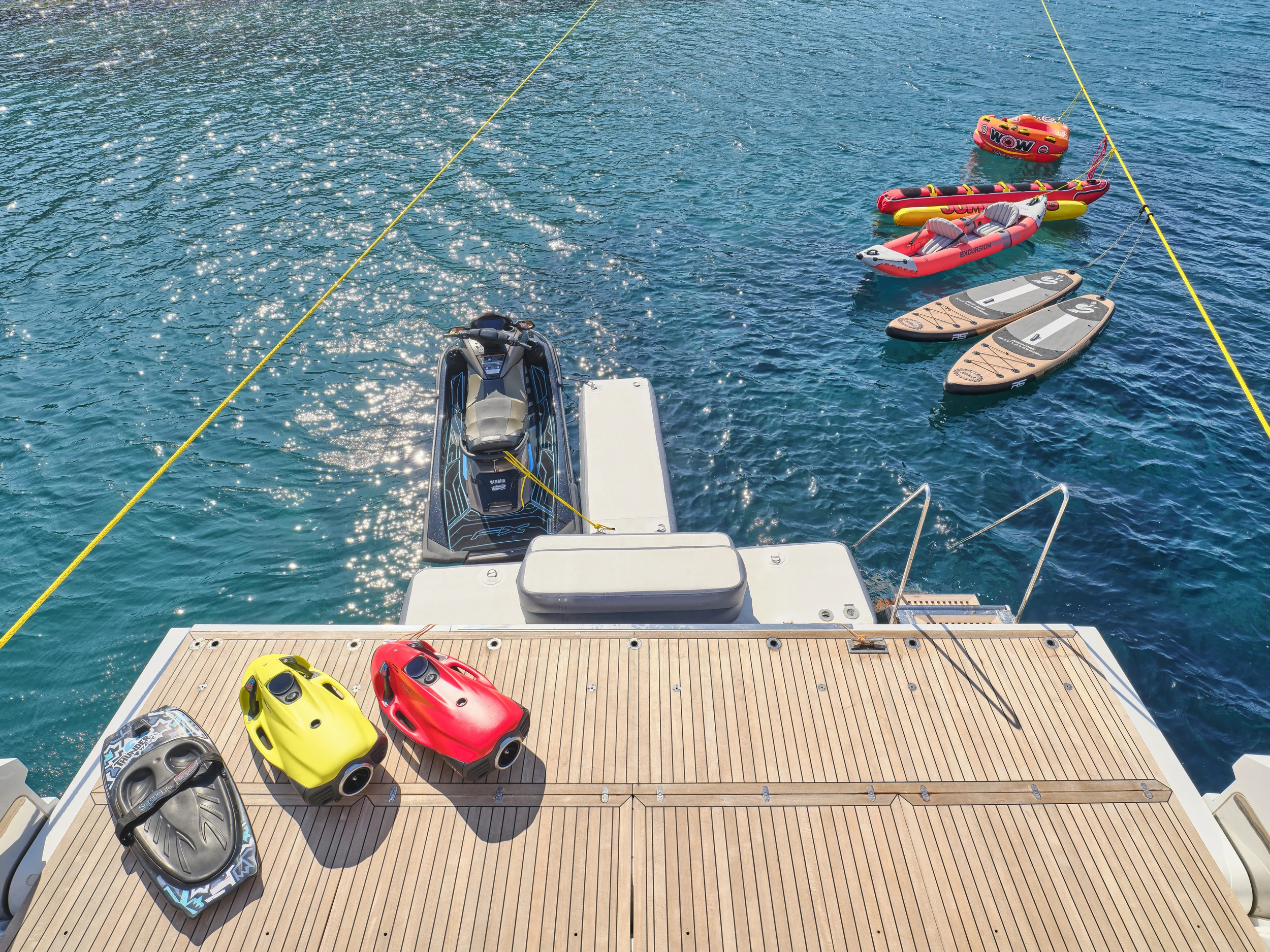 View from a boat showing a dock with three watercraft on the deck, a jet ski to the left and two remote-controlled boats in yellow and red, along with various inflated floatation devices and paddleboards floating in the water.