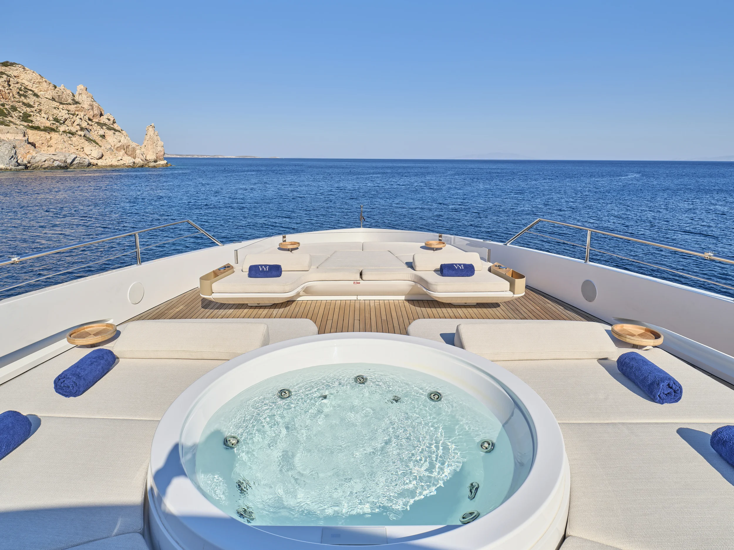 View from the deck of a yacht with a hot tub, surrounded by beige lounge chairs with blue towels, overlooking the ocean and rocky shoreline under a clear blue sky.