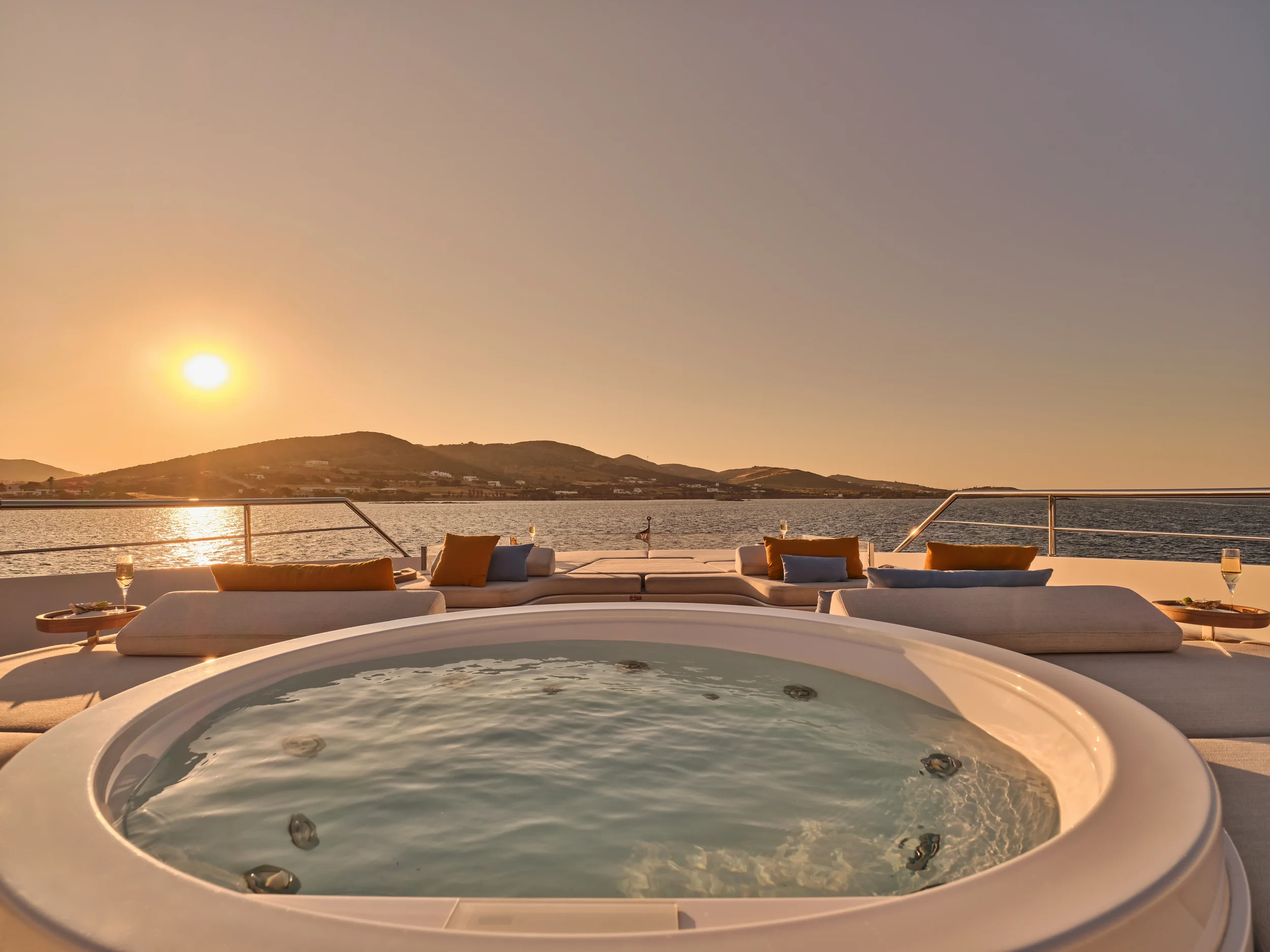 Luxurious outdoor hot tub on deck overlooking a body of water with mountains in the distance during sunset, with lounge chairs, cushions, and glasses of champagne nearby.