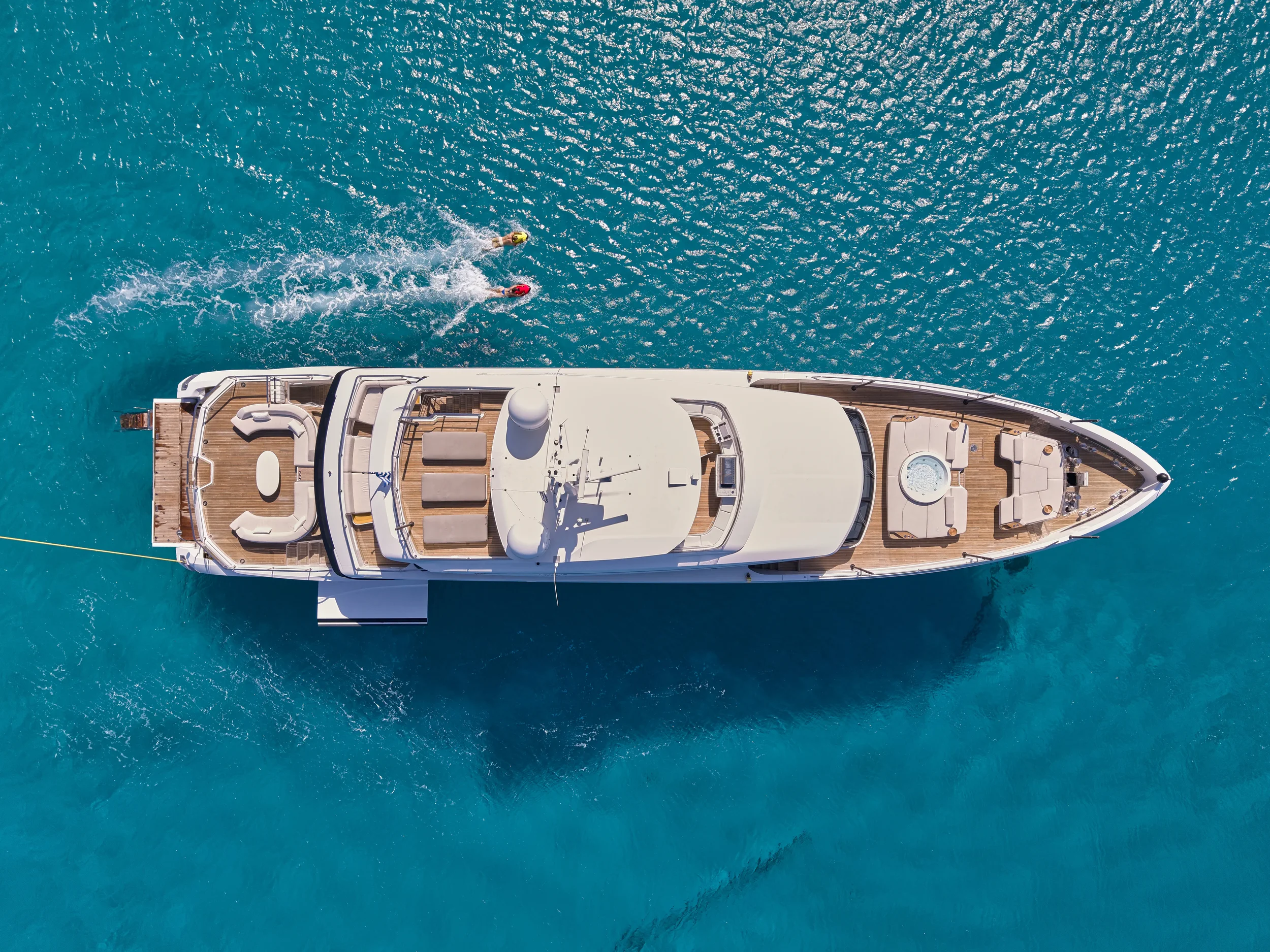Aerial view of a large white yacht sailing in clear blue water with two swimmers nearby.