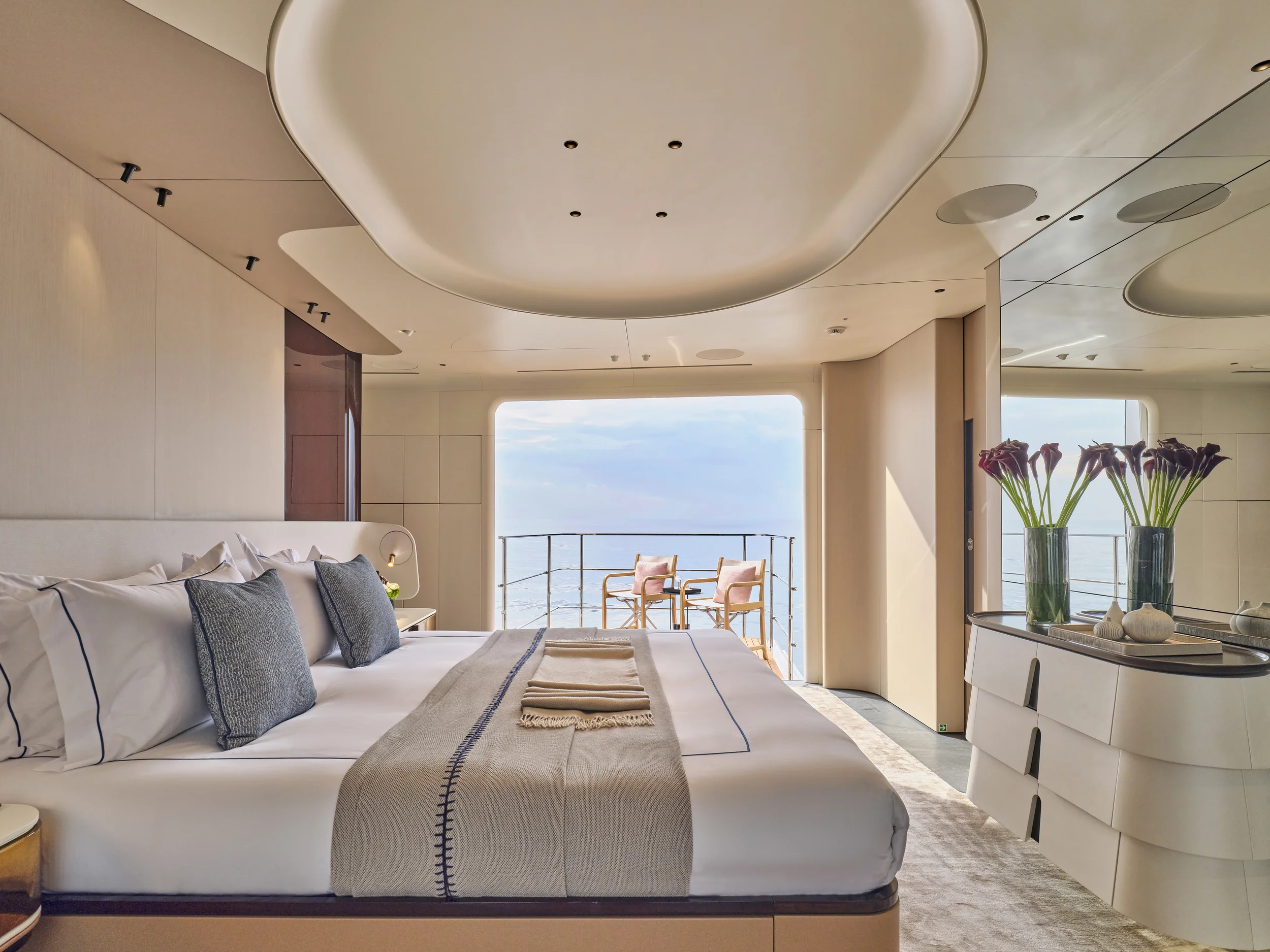 Sophisticated hotel room with a large bed, two chairs on a balcony overlooking the water, and modern decor including vases with flowers.