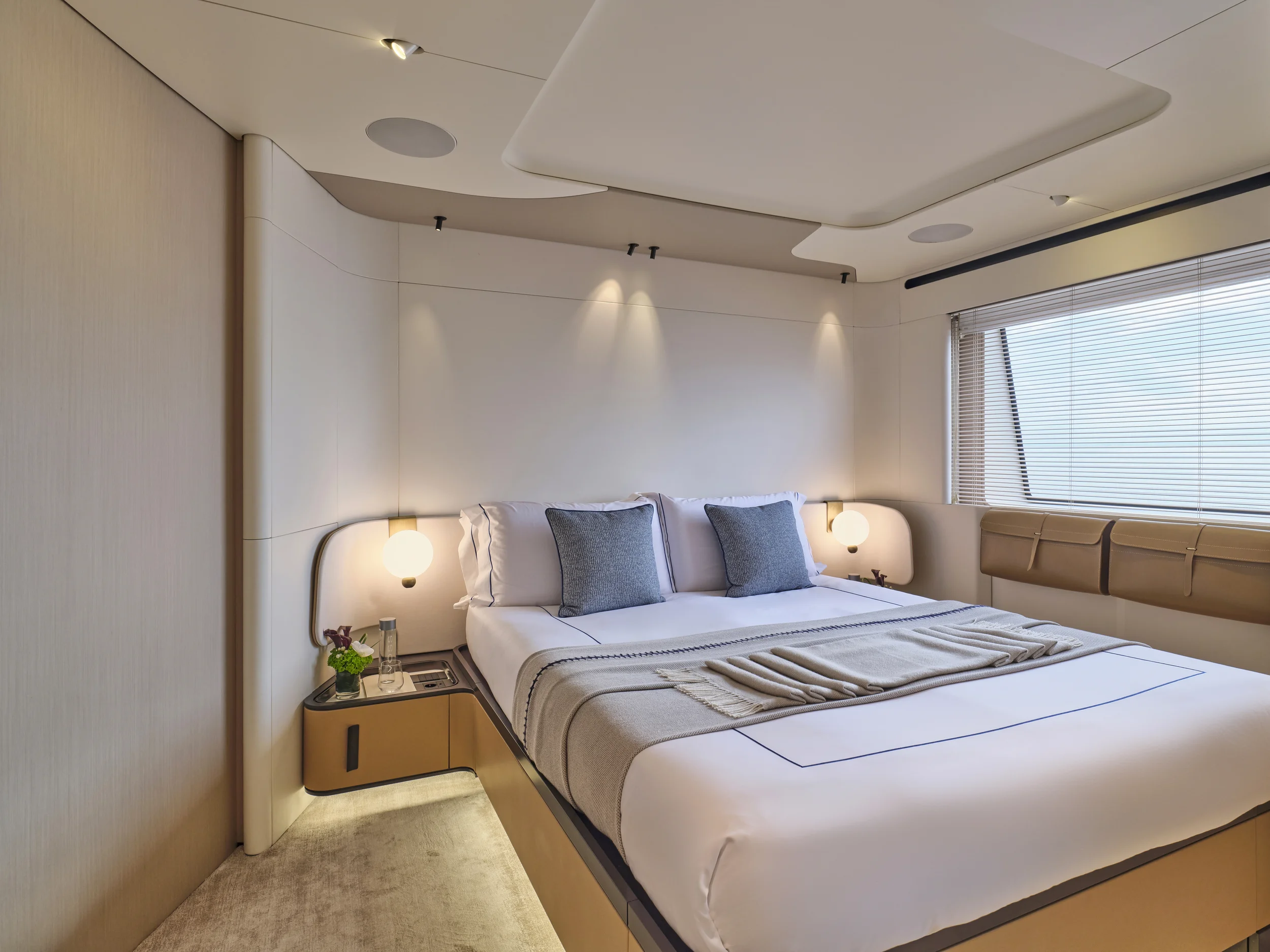 A modern hotel bedroom with a large bed, two gray pillows, bedside tables with lamps, a window with blinds, and beige accents.