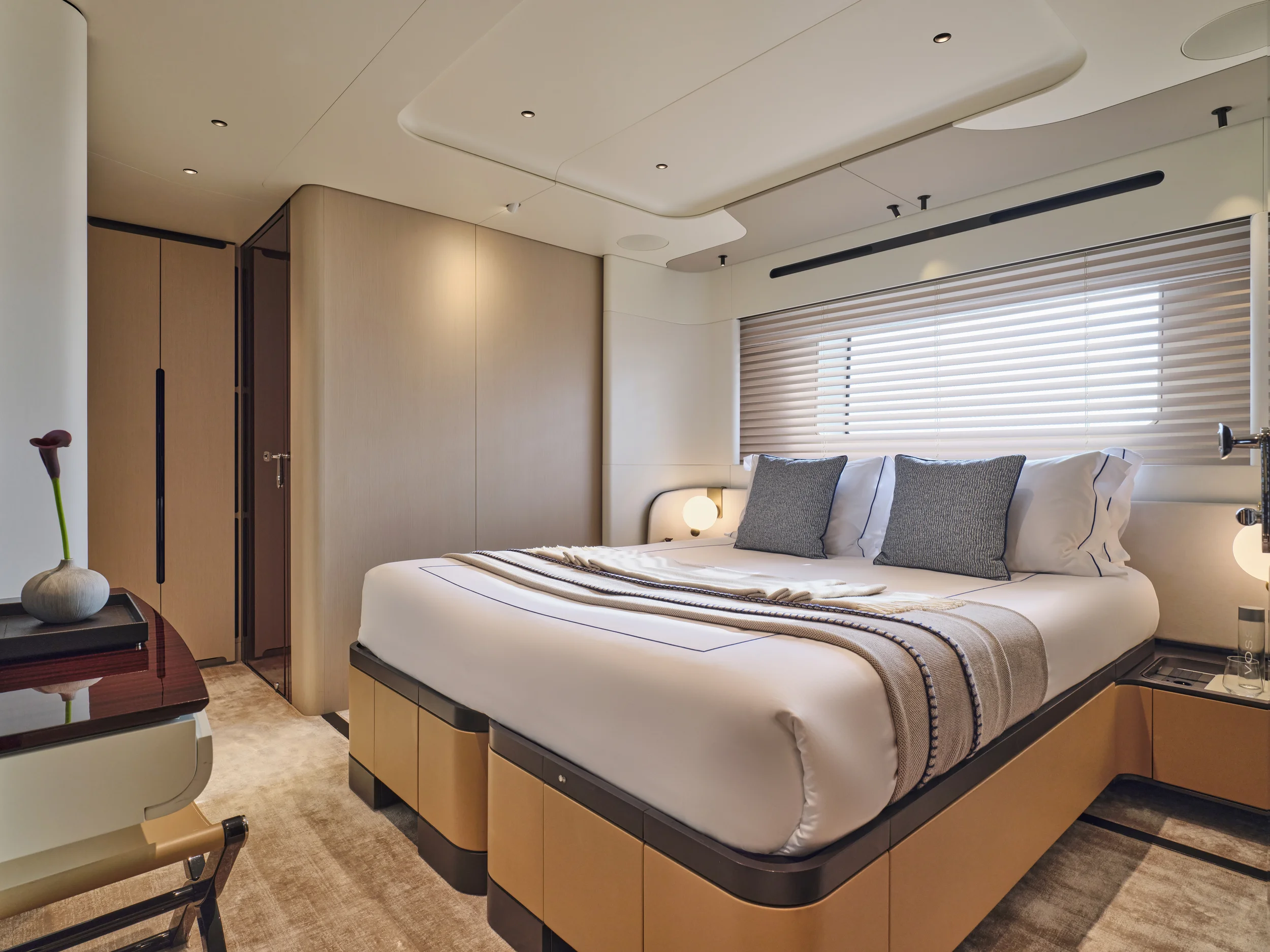 Modern hotel room with a large bed, two bedside tables with lamps, beige walls, and window with wooden blinds.