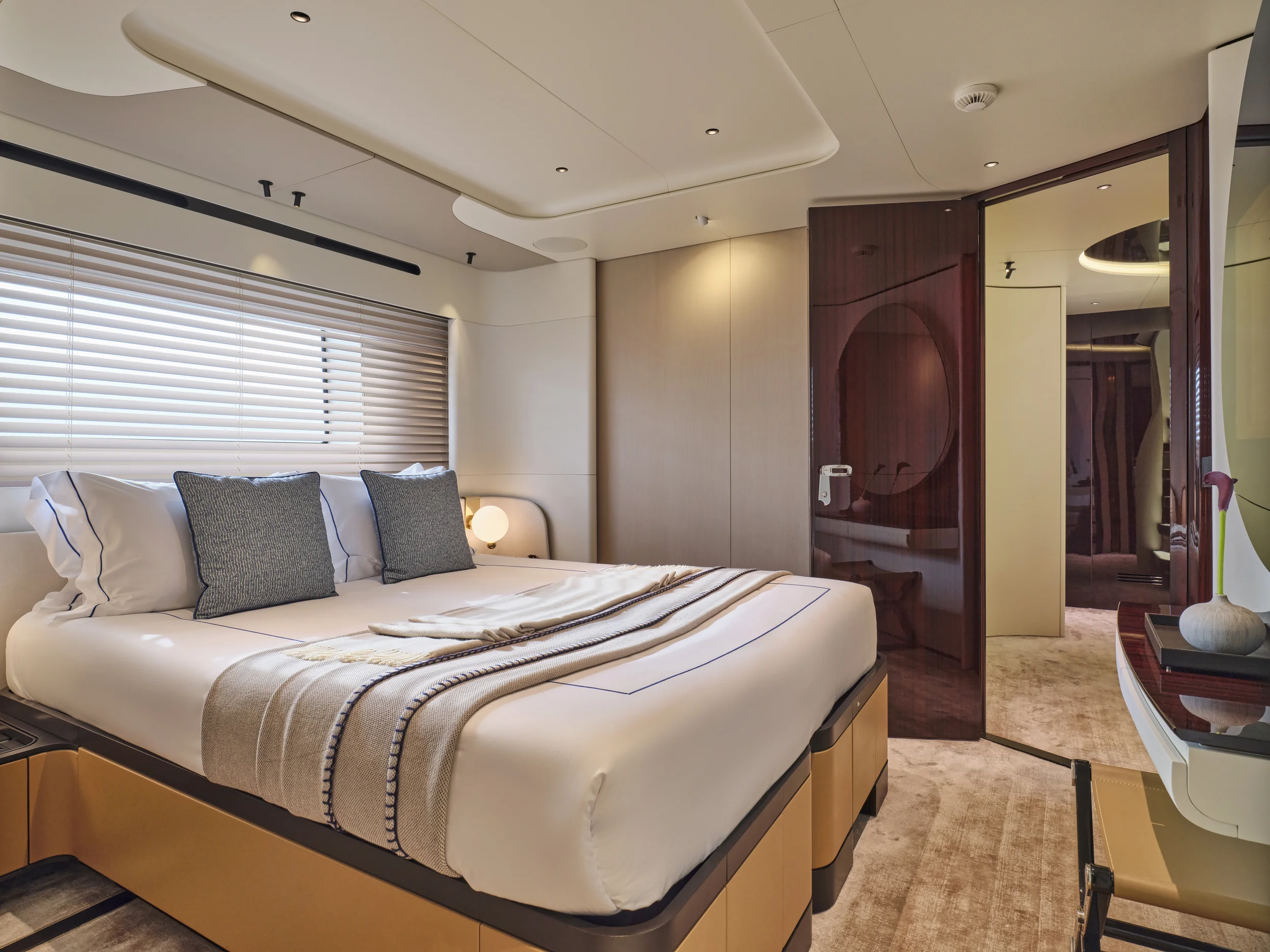 Luxurious bedroom with a large bed, gray pillows, and beige blanket, featuring a modern design with wooden accents, a window with horizontal blinds, and a door leading to another room.
